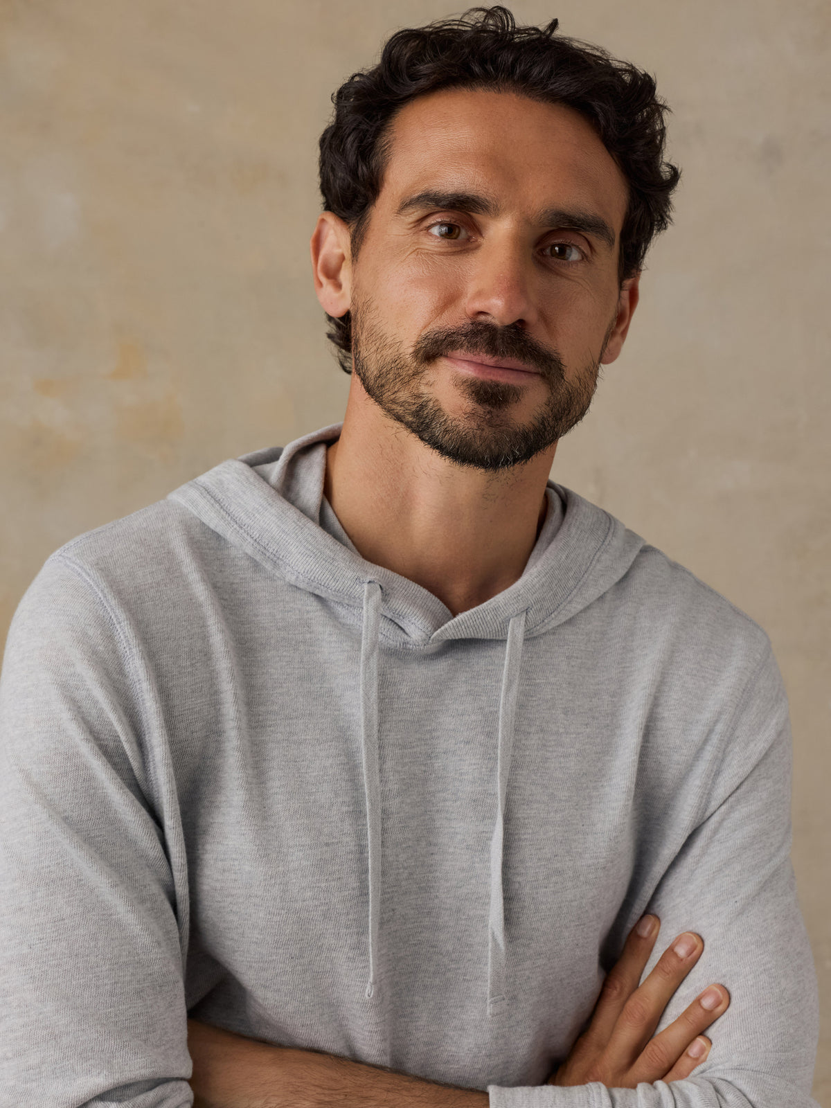 A man with dark curly hair and a beard wearing a light gray heathered pullover hoodie, standing with arms crossed against a textured beige background., image 2 of 8