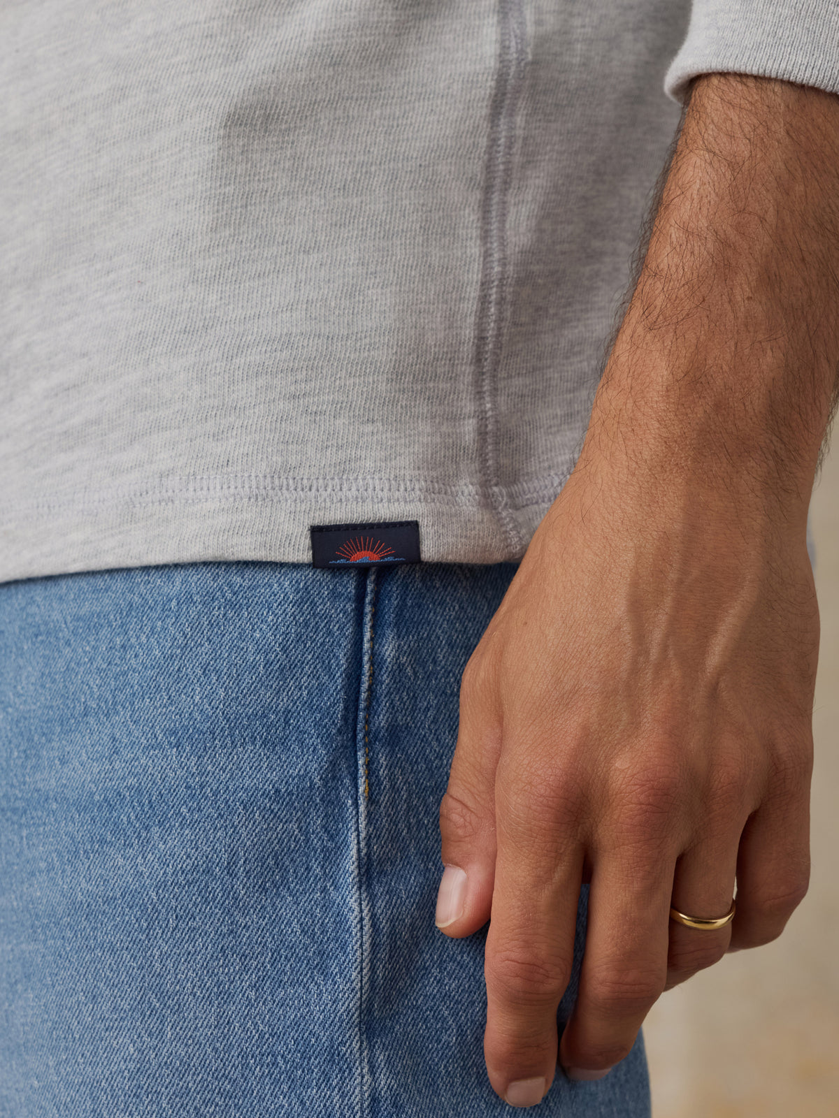 Close-up of a man wearing a light gray heathered knit top and blue jeans, with a small dark waistband label visible and a gold ring on the finger., image 4 of 8