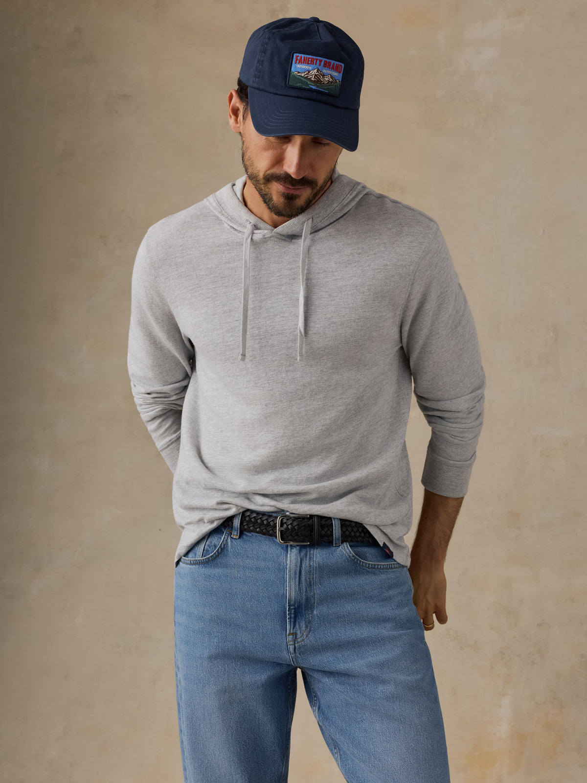 A man wearing a light gray long-sleeve hoodie, blue jeans, a black belt, and a navy blue baseball cap with a Patagonia logo patch, standing against a textured beige wall., image 6 of 8