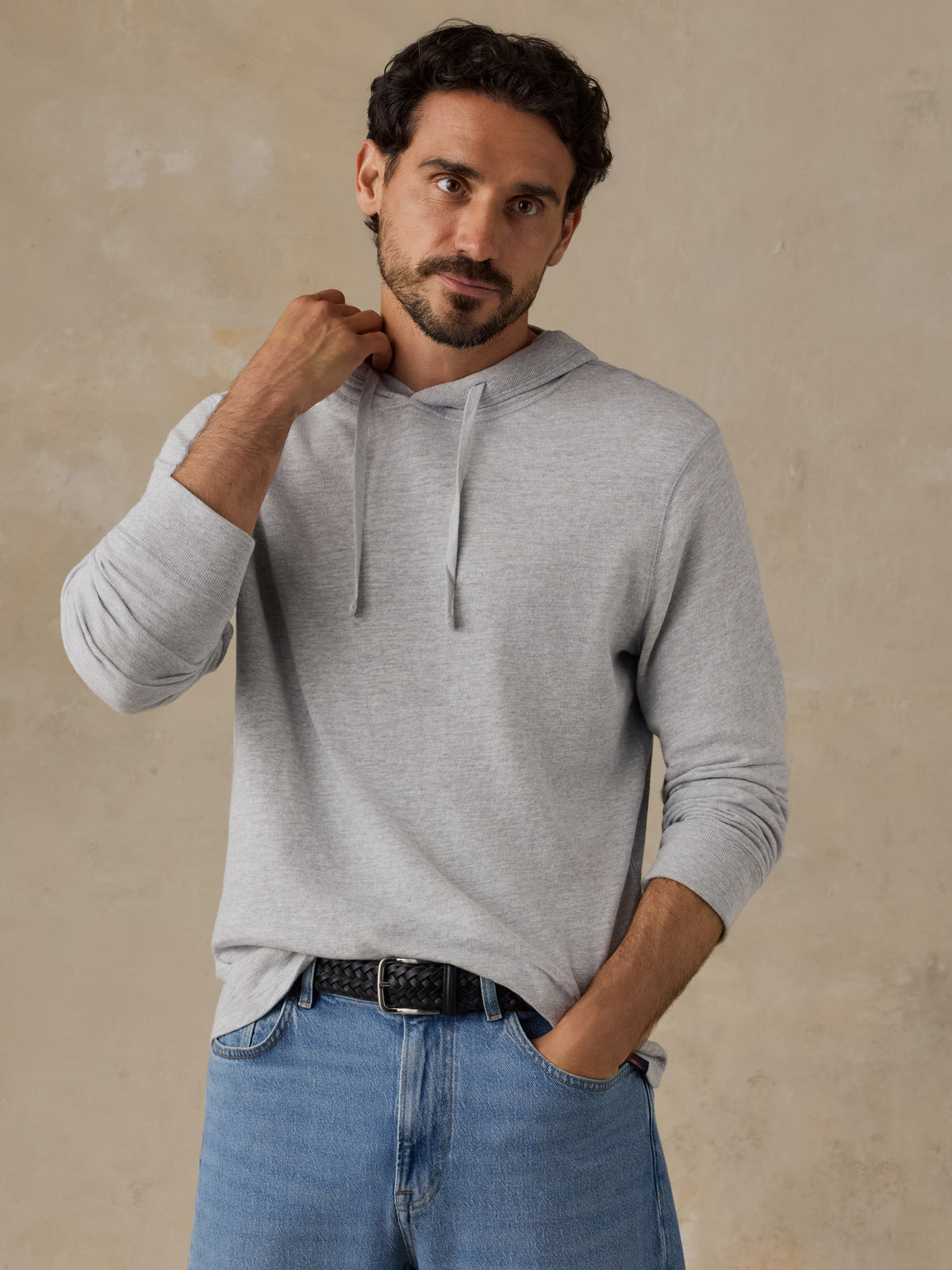 A man wearing a light gray long-sleeve hoodie with drawstrings, paired with blue jeans and a black belt, standing against a textured beige wall., image 1 of 8