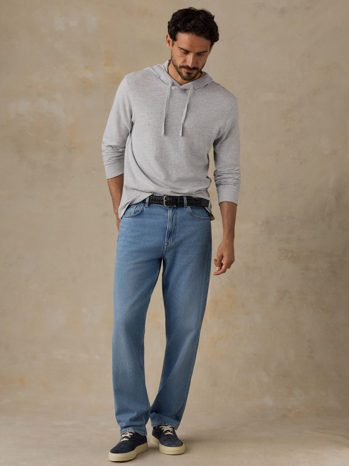 A man wearing a light gray long-sleeve hoodie, blue jeans, a black belt, and navy sneakers, standing against a textured beige studio backdrop., image 5 of 8