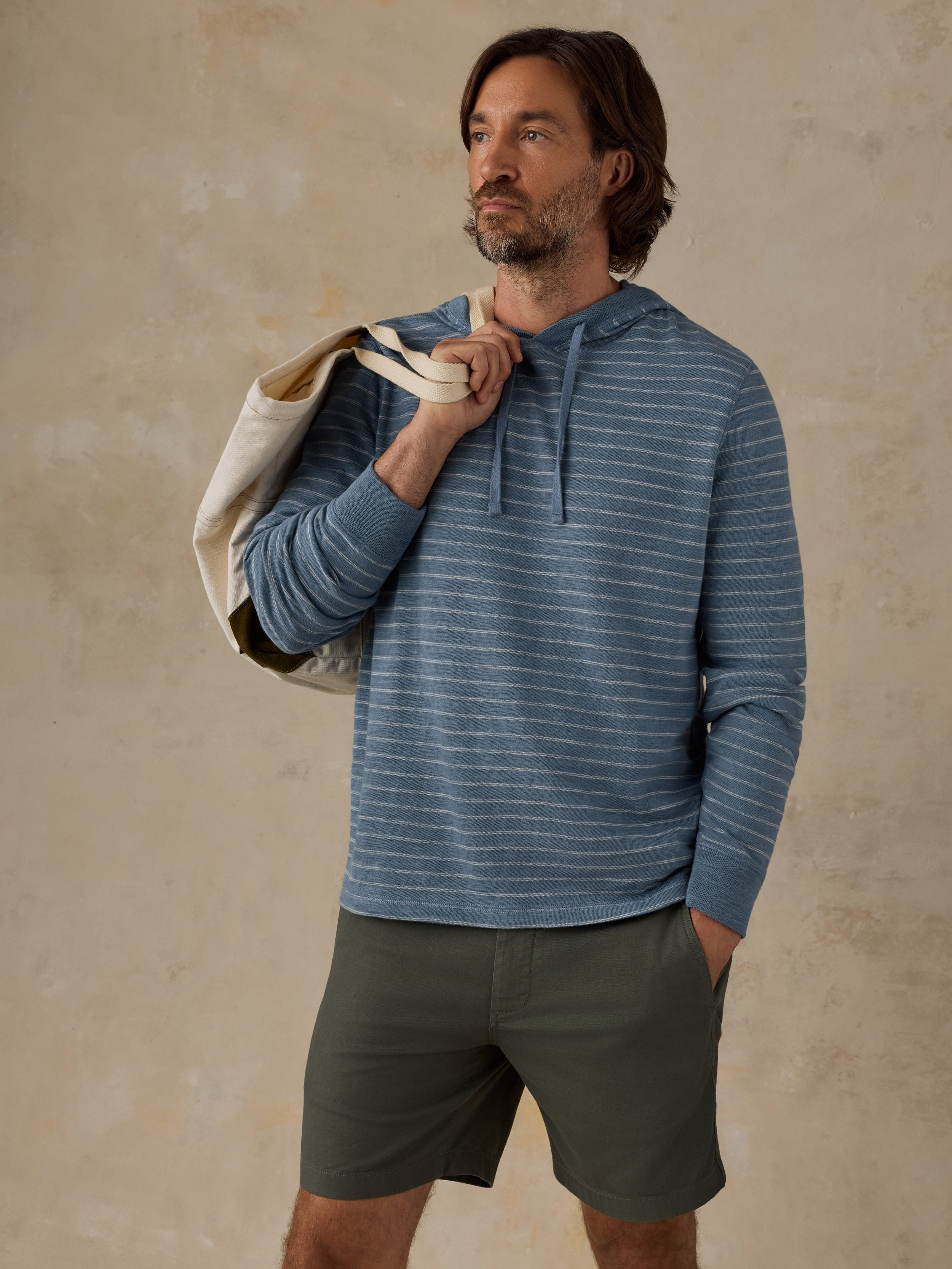 A man wearing a blue and white striped long-sleeve hoodie and olive green shorts, holding a cream canvas tote bag over his shoulder against a textured beige studio backdrop.