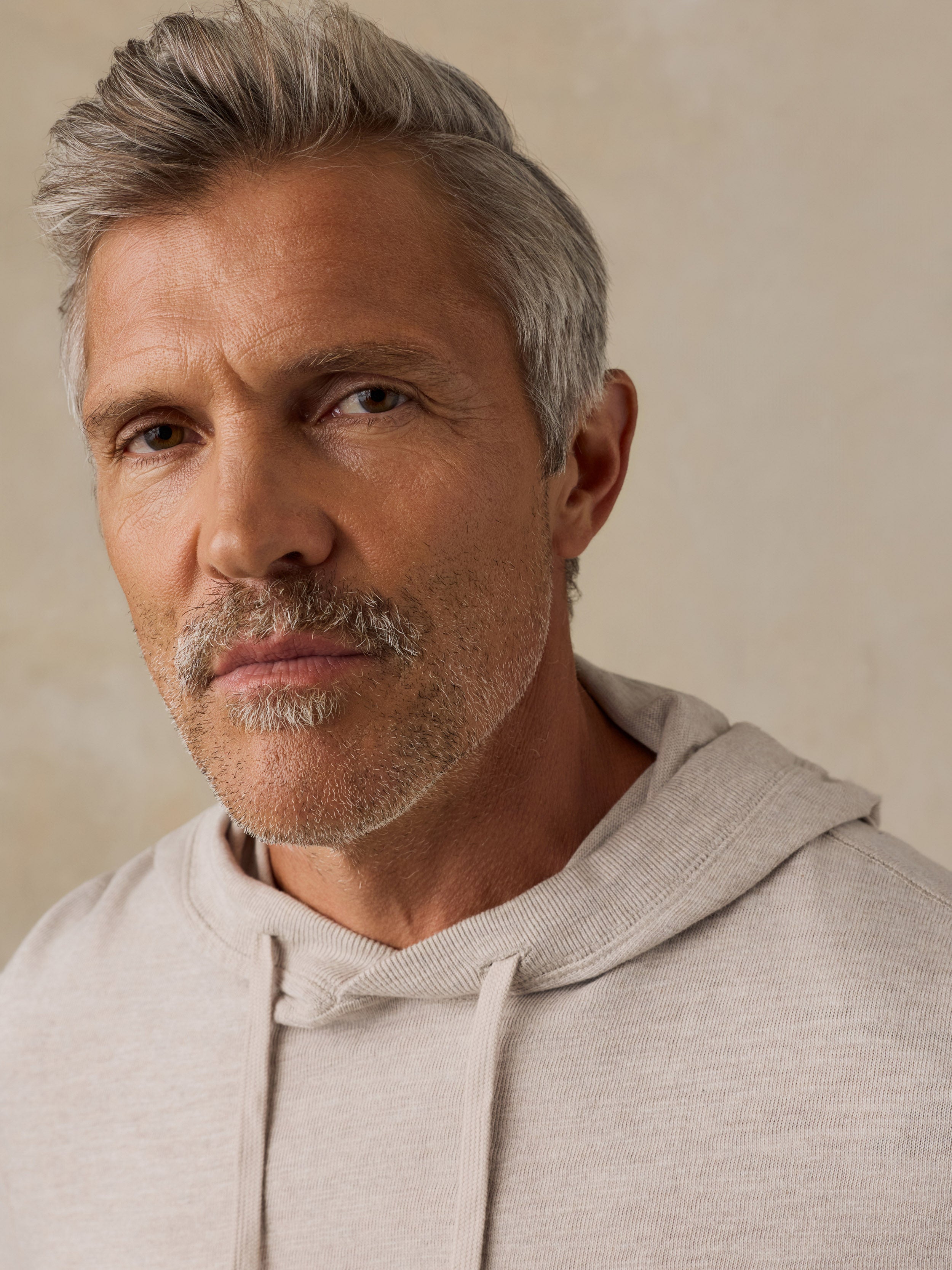 A close-up portrait of a man with gray hair and a mustache wearing a light beige hooded sweatshirt, set against a neutral beige background.
