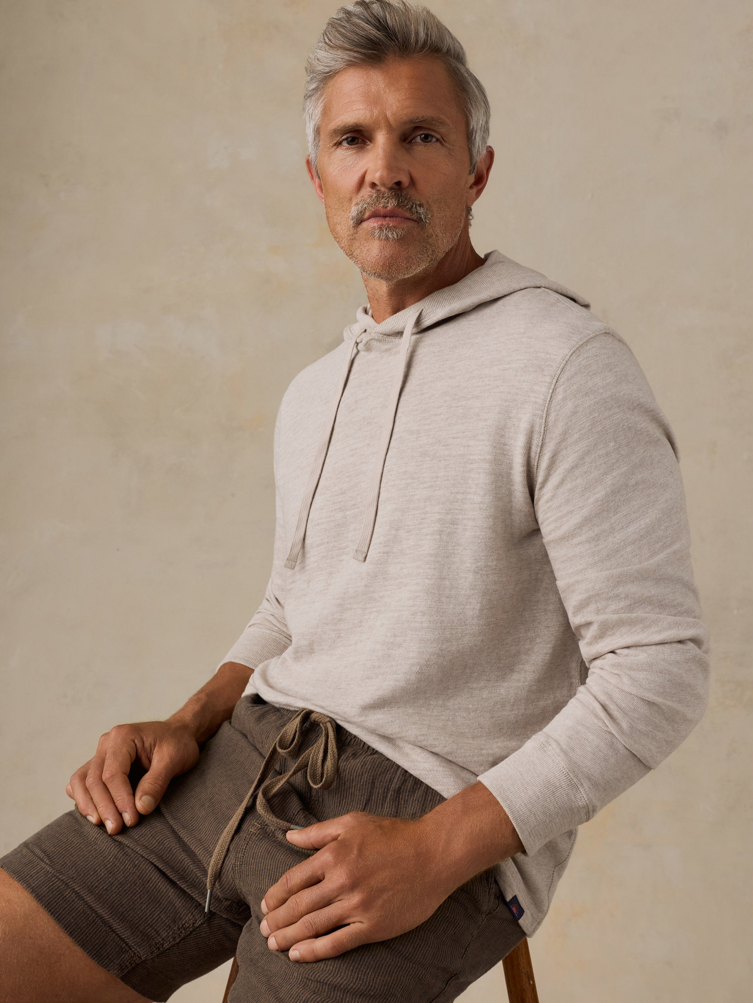 A man wearing a light beige long-sleeve hooded sweatshirt and brown drawstring shorts, seated against a textured beige studio background.