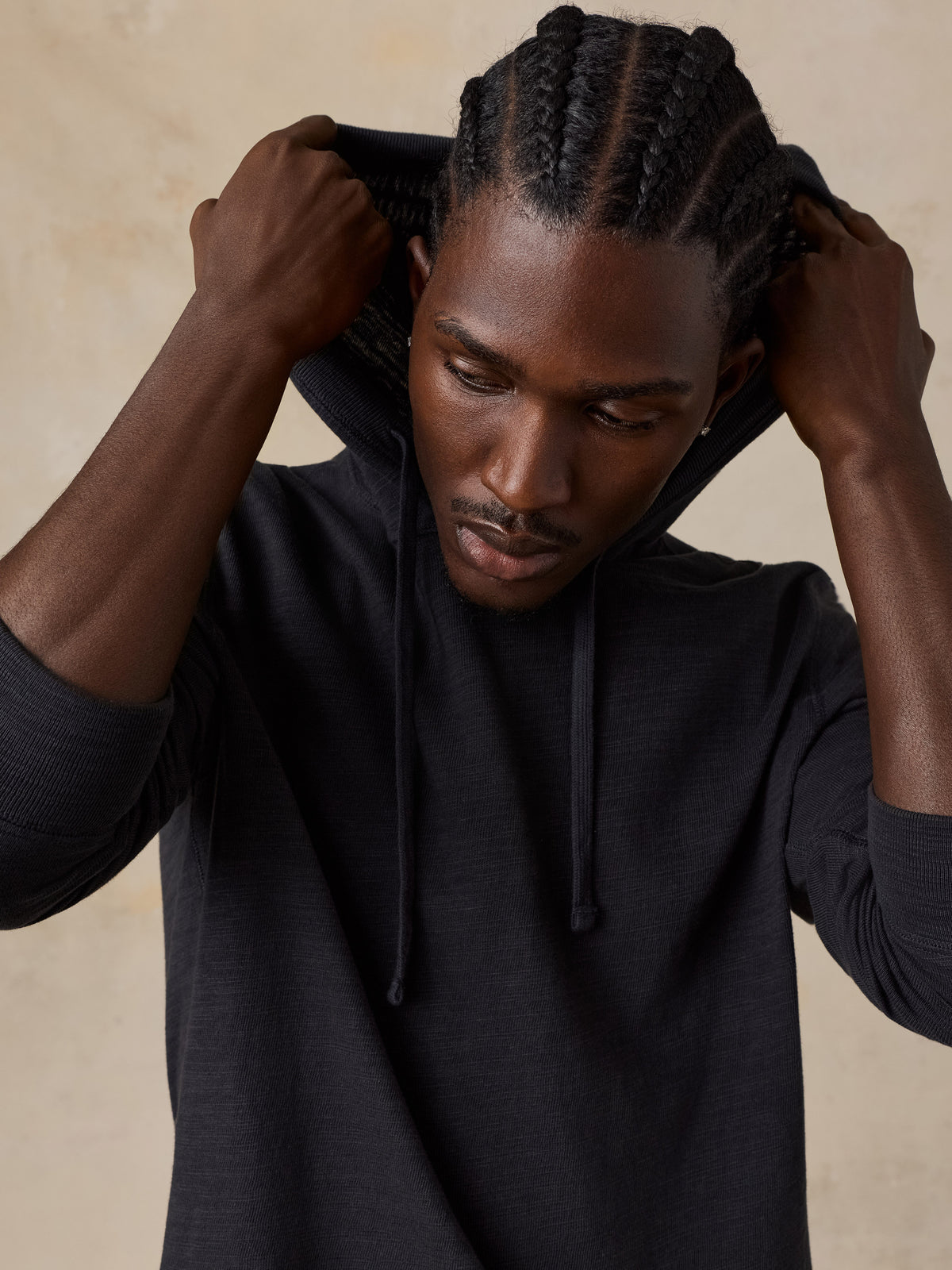 Man wearing a black hoodie over a blue t-shirt against a beige studio background, image 4 of 7