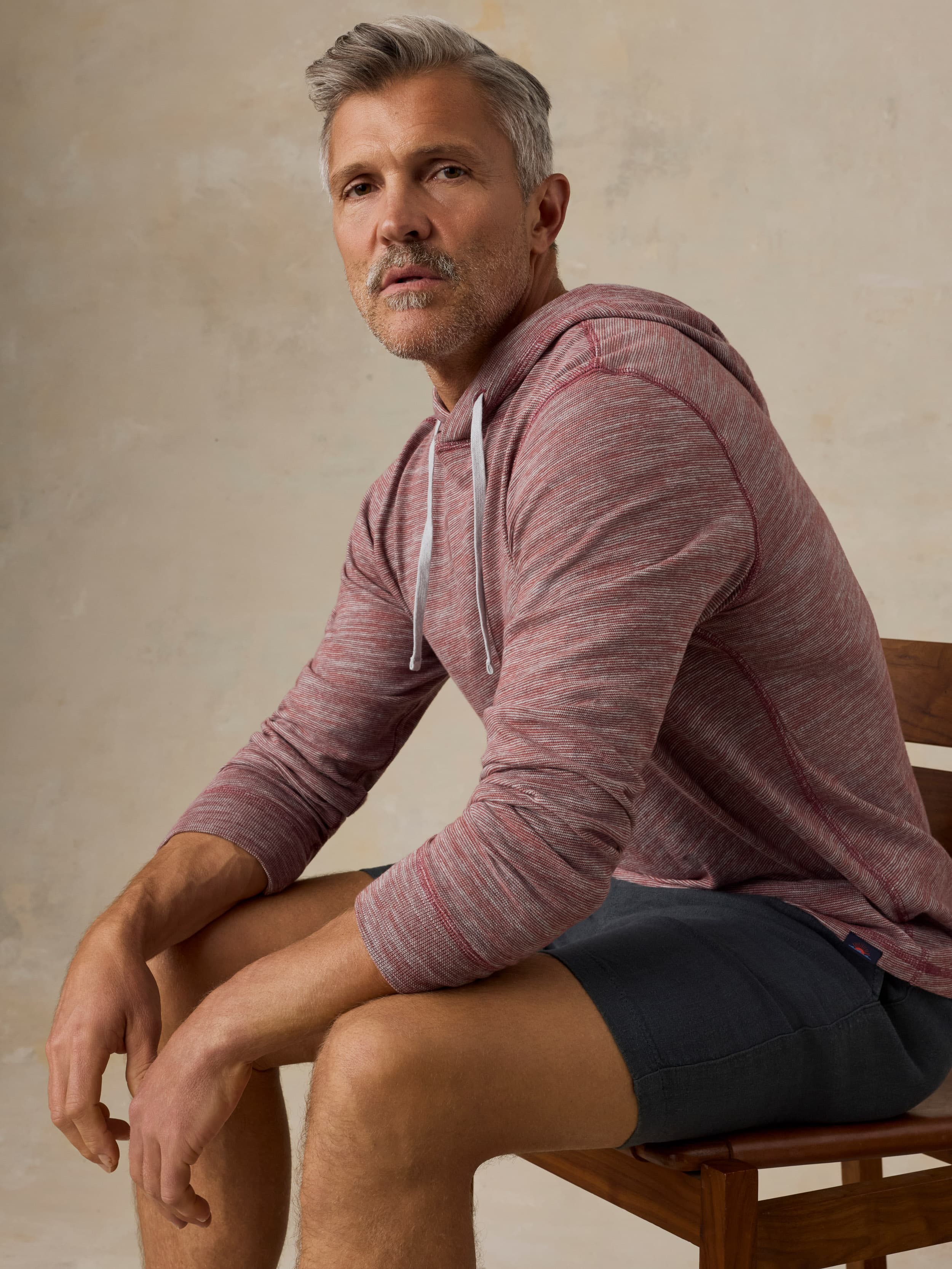 Man sitting on a wooden chair wearing a red heathered hoodie and black shorts against a beige background