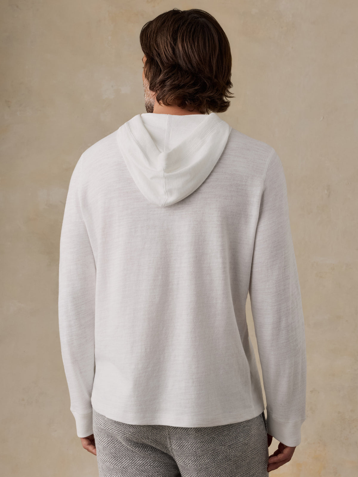A man viewed from behind wearing a light off-white hoodie and gray textured pants, standing against a warm beige plaster wall., image 4 of 8