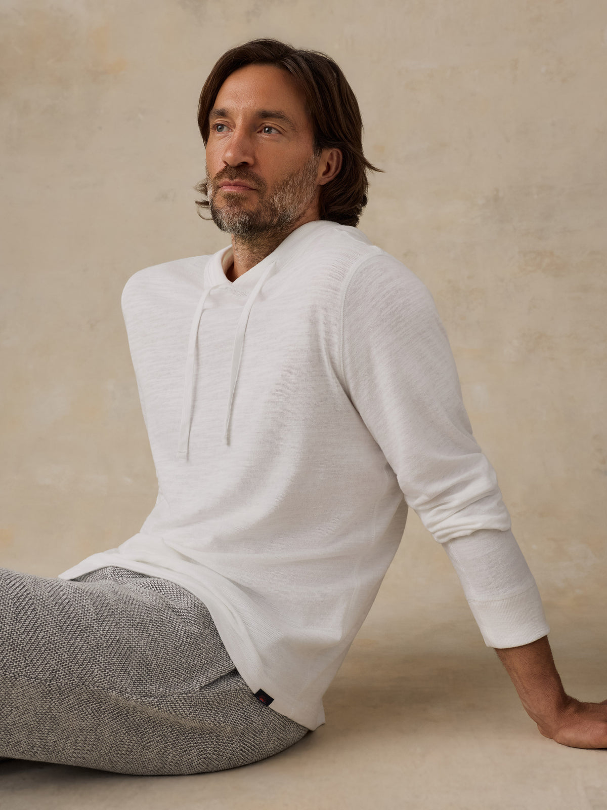 A man wearing a white long-sleeve hoodie with a drawstring neckline and gray textured trousers, seated against a warm beige plaster wall., image 2 of 8