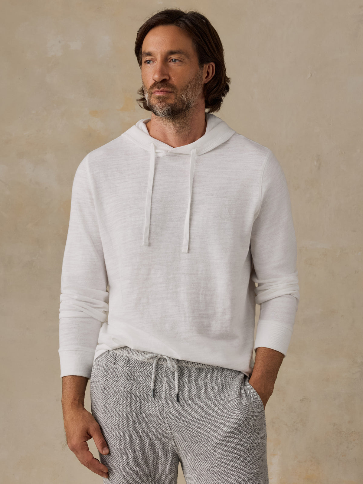 A man wearing a light off-white long-sleeve hoodie and heather gray drawstring sweatpants, standing against a textured beige wall., image 1 of 8