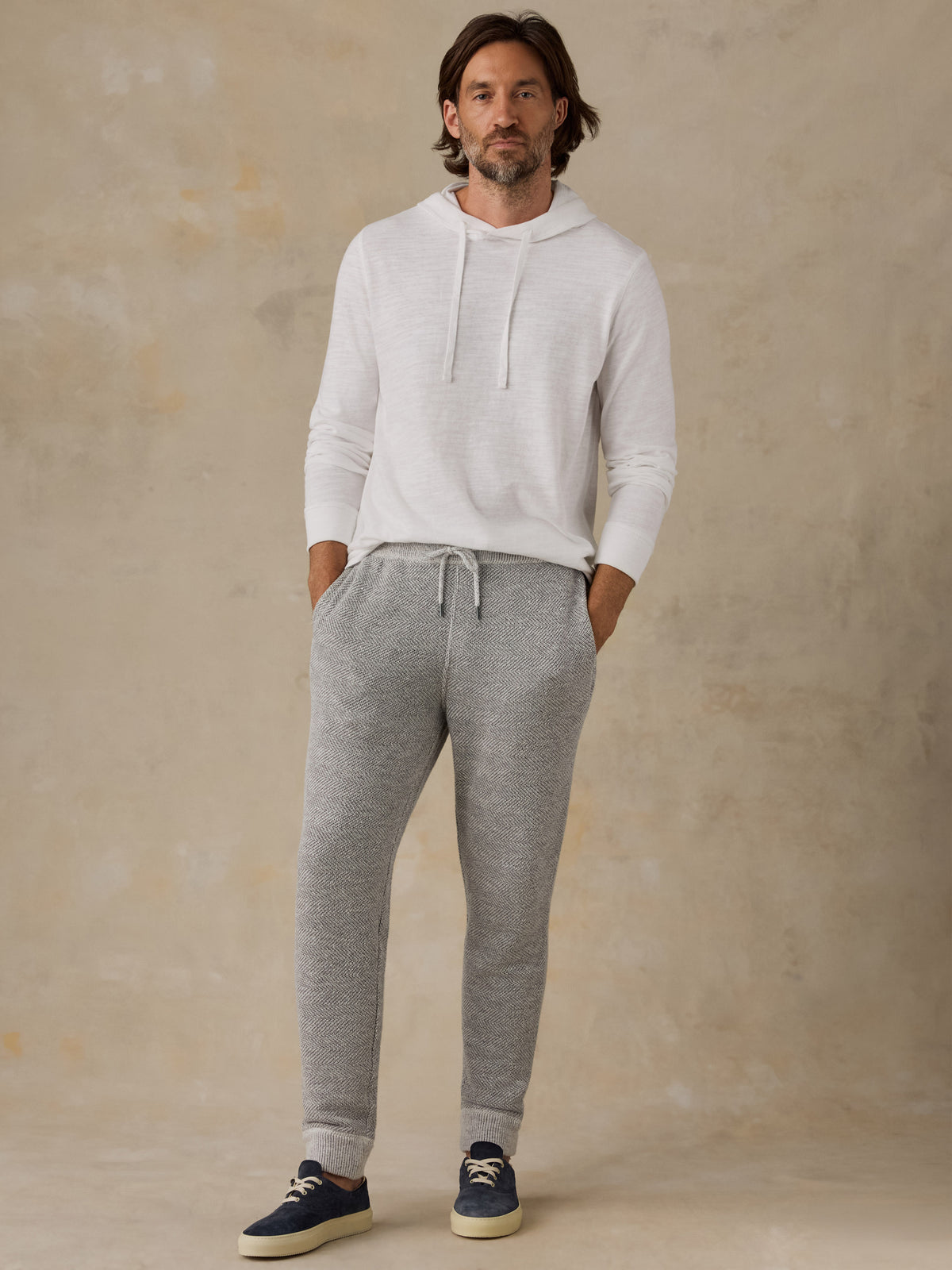 A man wearing a white long-sleeve hoodie and heather gray jogger sweatpants, standing against a textured beige studio backdrop., image 6 of 8