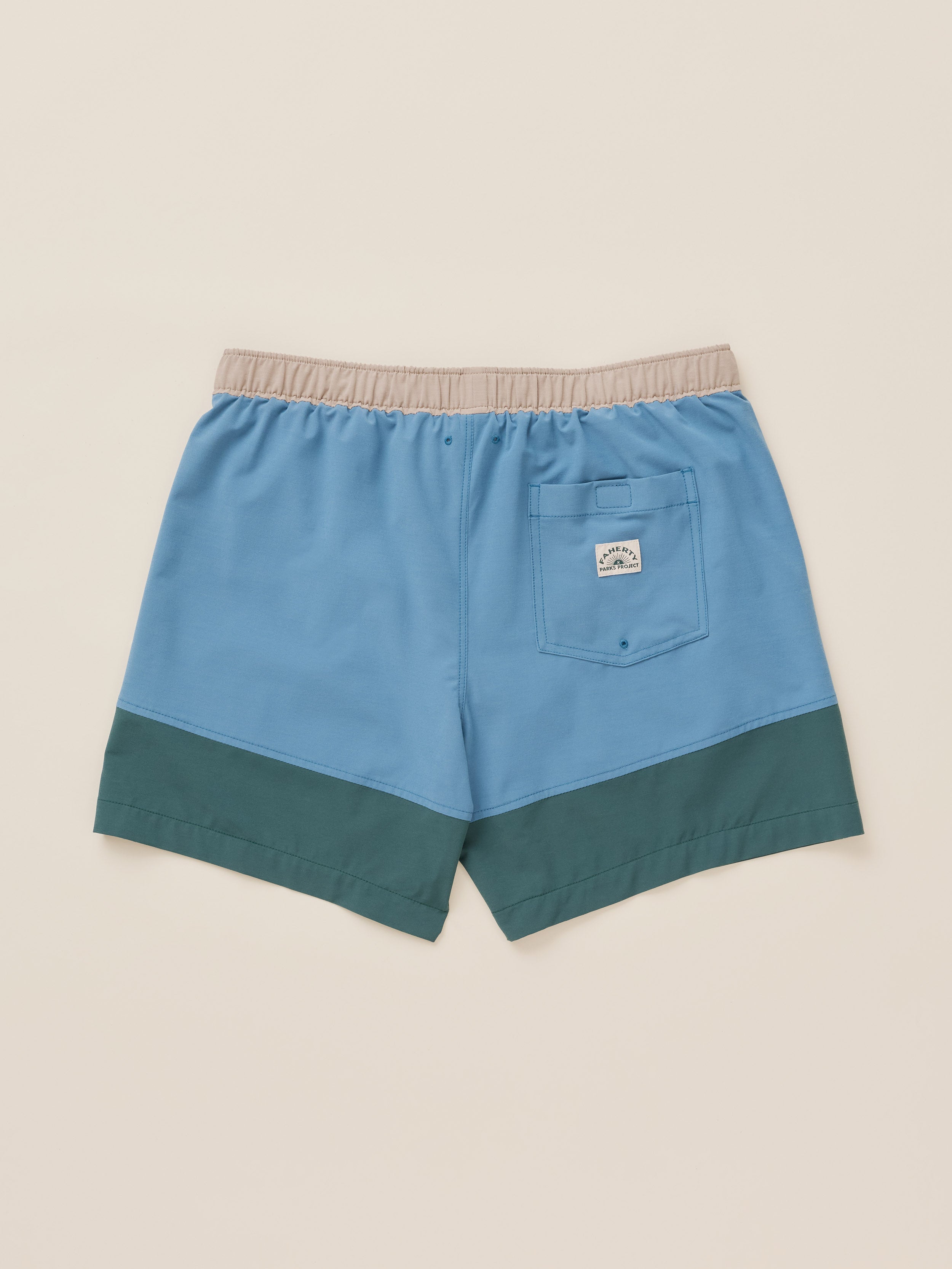 A pair of color-block swim trunks shown from the back, featuring a light blue upper body, a dark teal lower hem, and a beige elastic waistband. A small rectangular Faherty brand patch is visible on the back pocket.