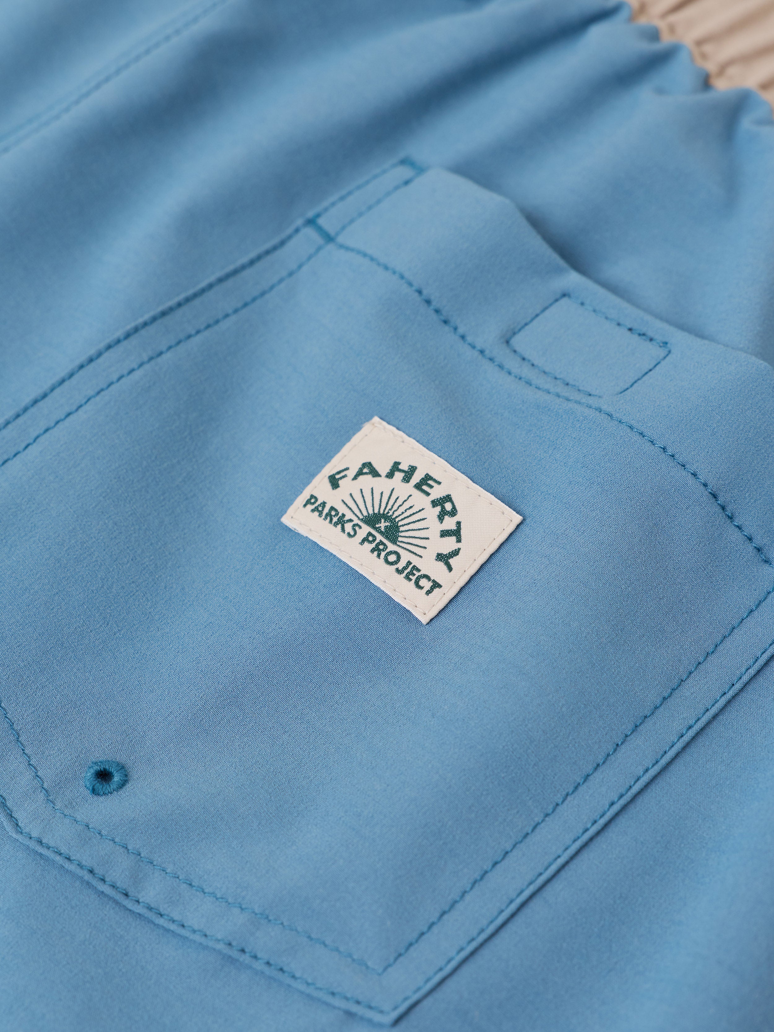 A close-up of a light blue fabric garment, likely shorts or pants, showing a front pocket with a white sewn-in label that reads 'FAHERTY x PARKS PROJECT'.