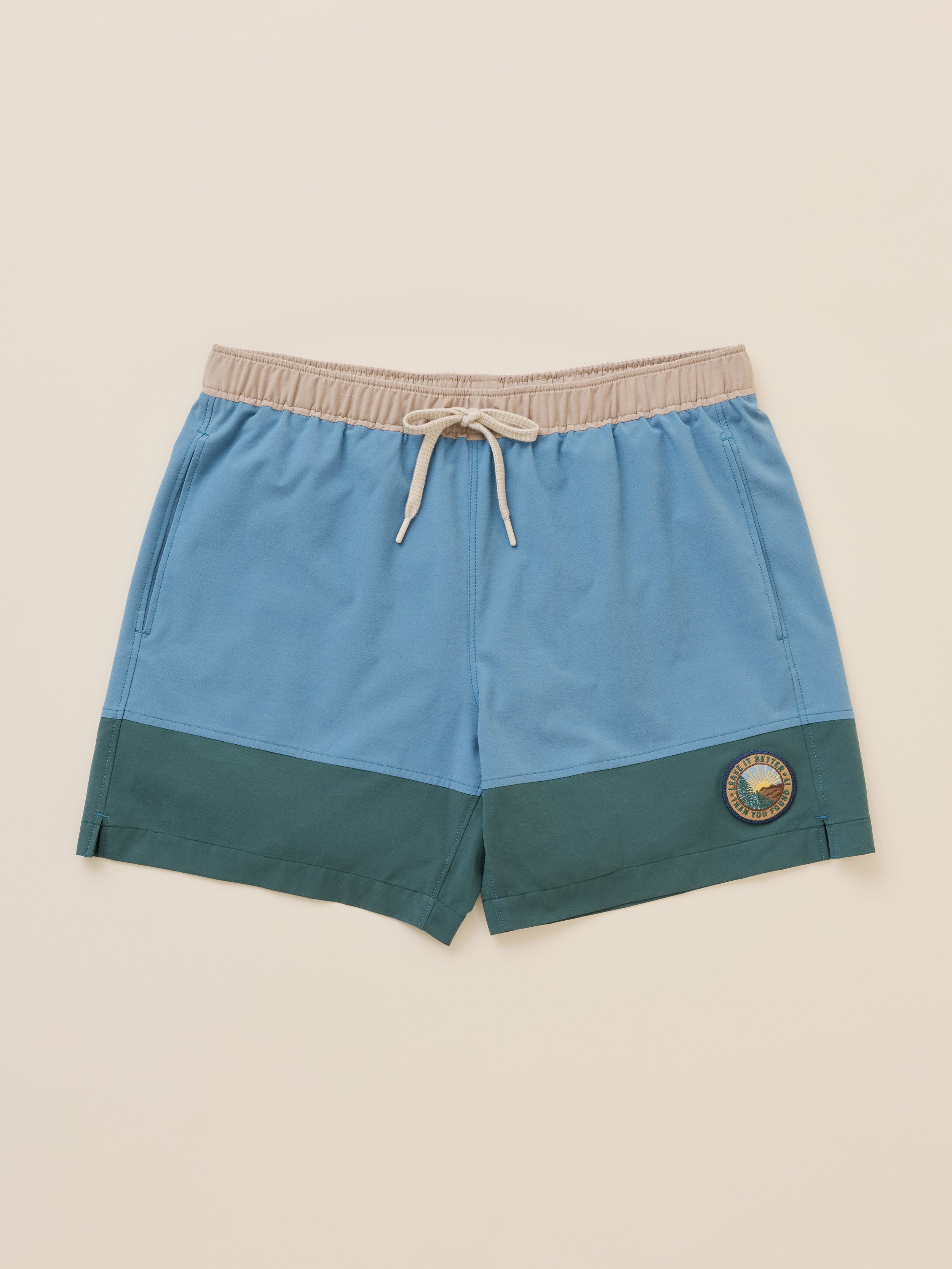 Colorblock swim shorts in light blue and dark teal green with a beige elastic waistband and white drawstring, featuring a circular logo patch on the lower left leg, shown flat on a warm cream background.