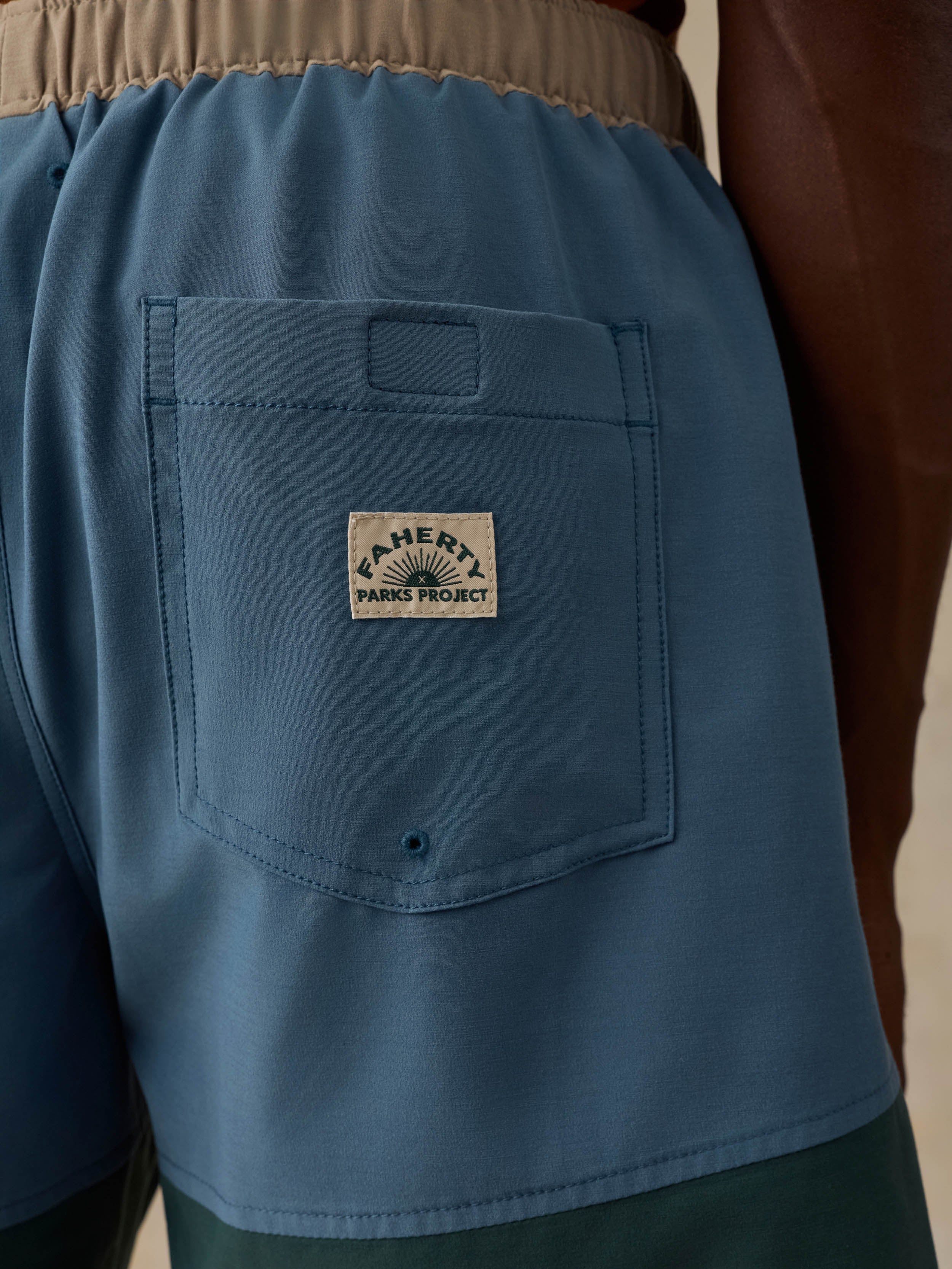 Close-up of a man wearing blue board shorts, showing the back pocket with a visible Parkway 'Parks Project' patch and a beige elastic waistband.