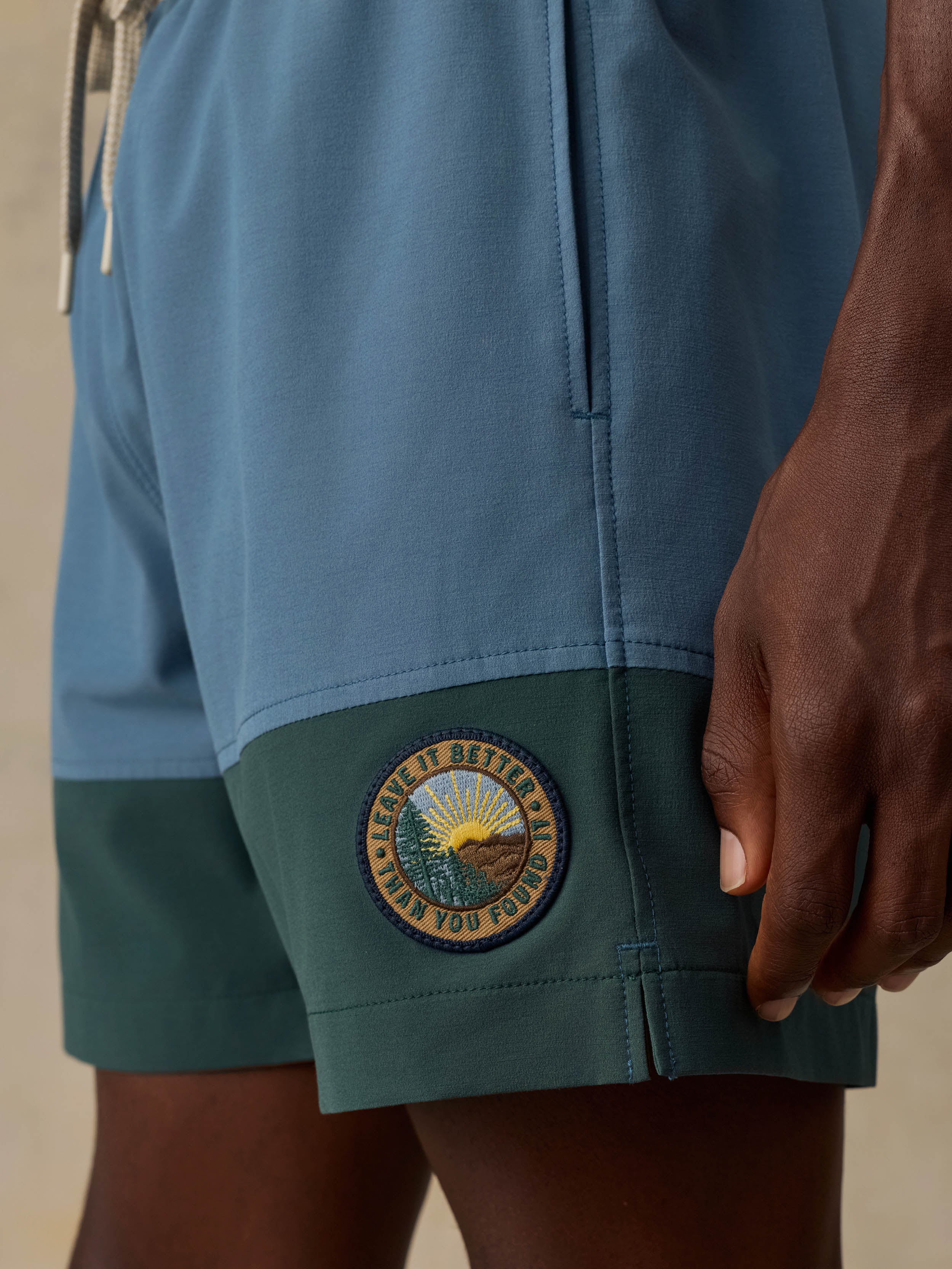 Close-up of a man wearing color-block board shorts in dusty blue and teal, with a circular embroidered patch on the lower left leg.