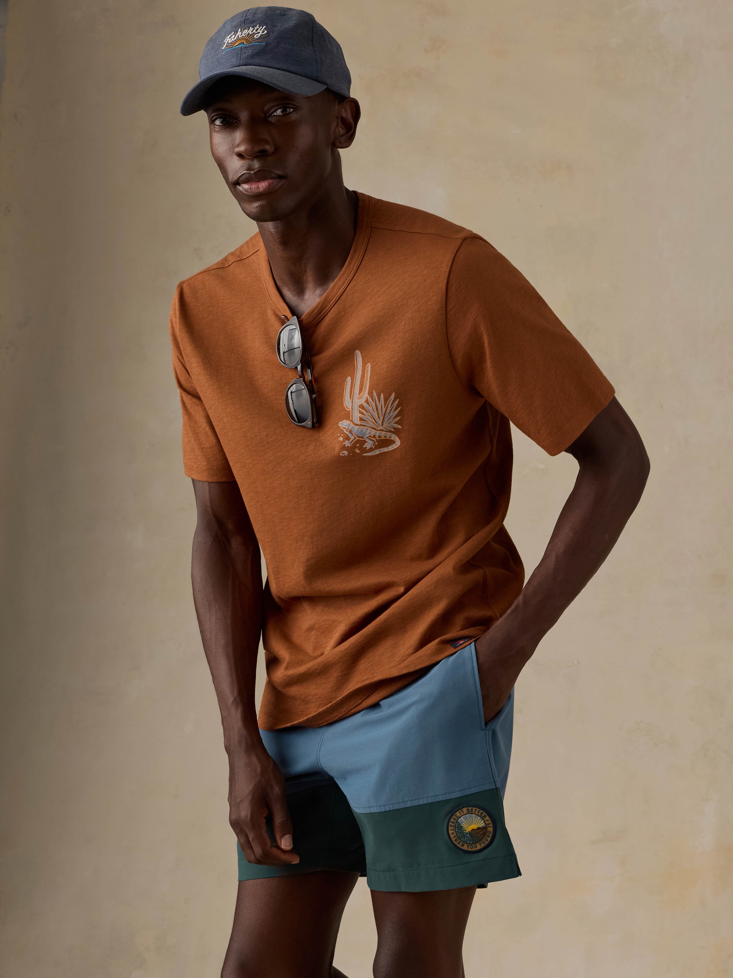 A man wearing a rust-orange t-shirt with a desert graphic, blue shorts, and a blue cap, posed against a textured beige background.