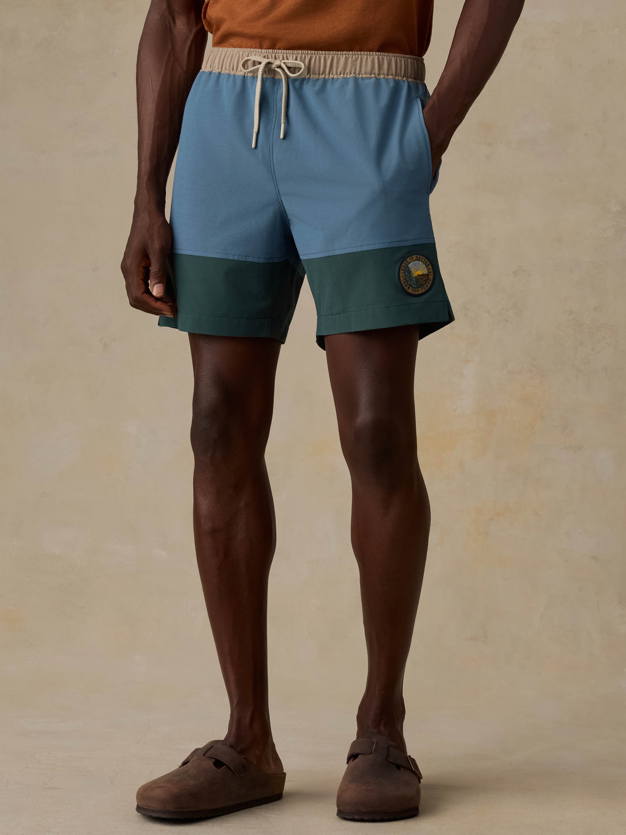 A man wearing colorblock swim shorts in light blue, dark teal, and beige, paired with a brown t-shirt and brown slip-on shoes, standing against a textured beige background.