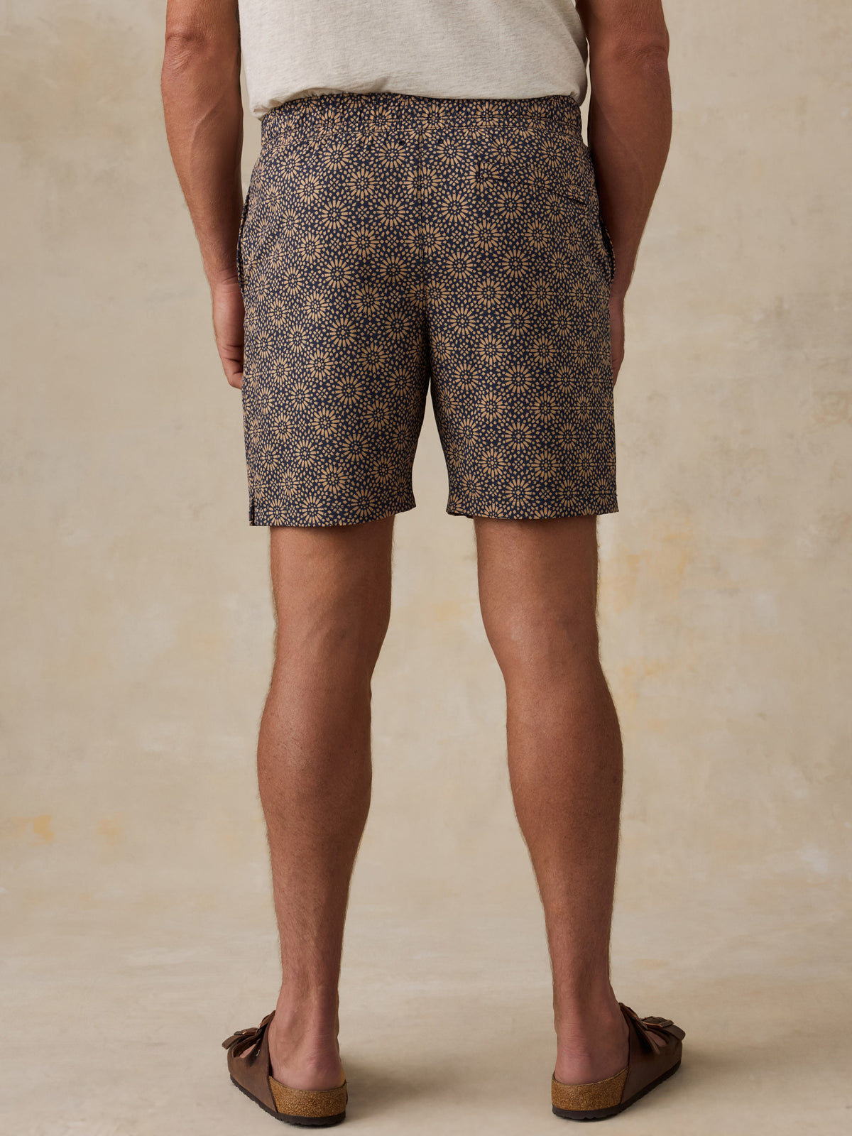 Man wearing a light colored t-shirt, brown patterned swim shorts, and sandals against a beige background, image 3 of 6