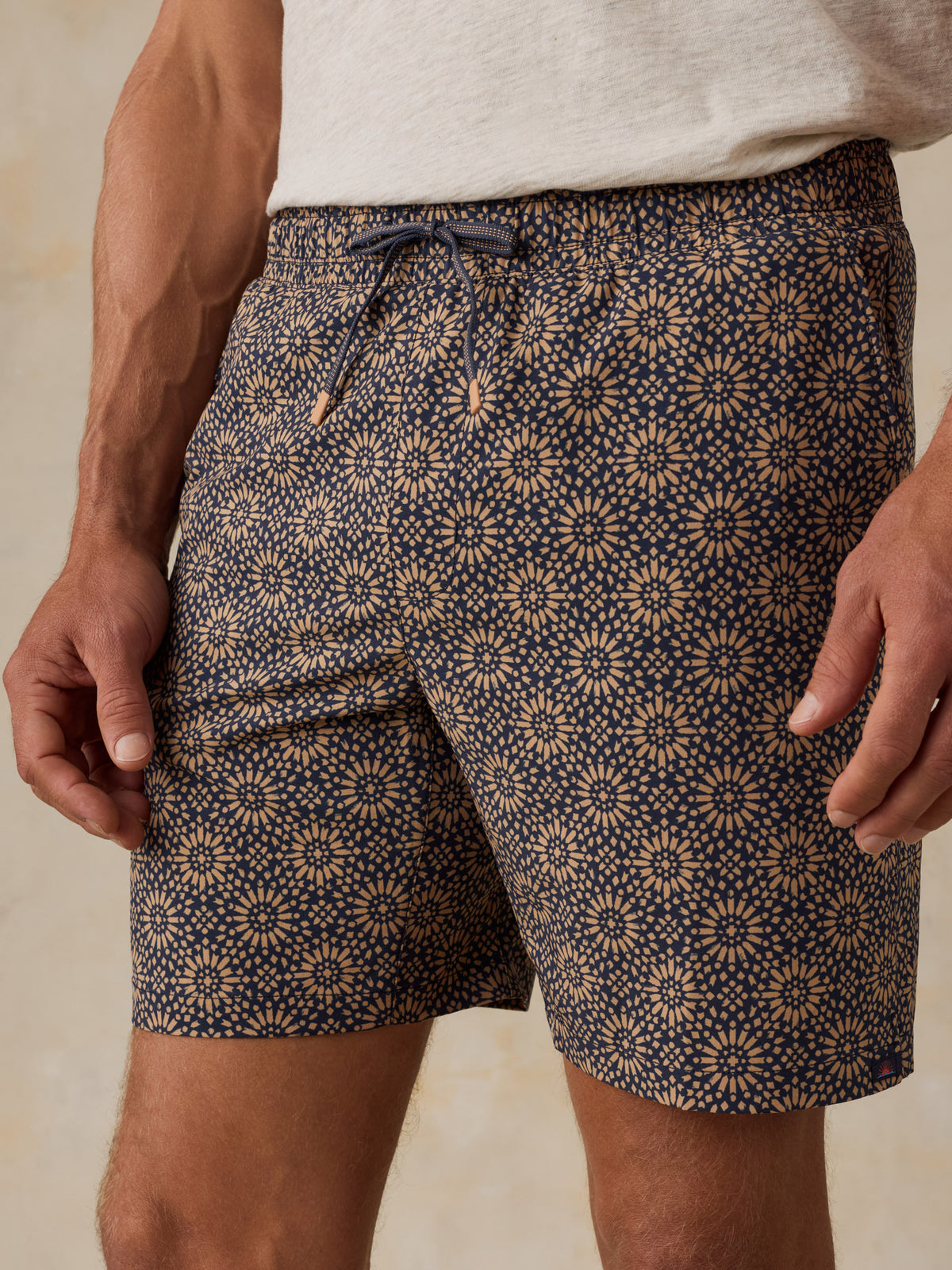 Man wearing a light colored t-shirt, brown patterned swim shorts, and sandals against a beige background, image 6 of 6