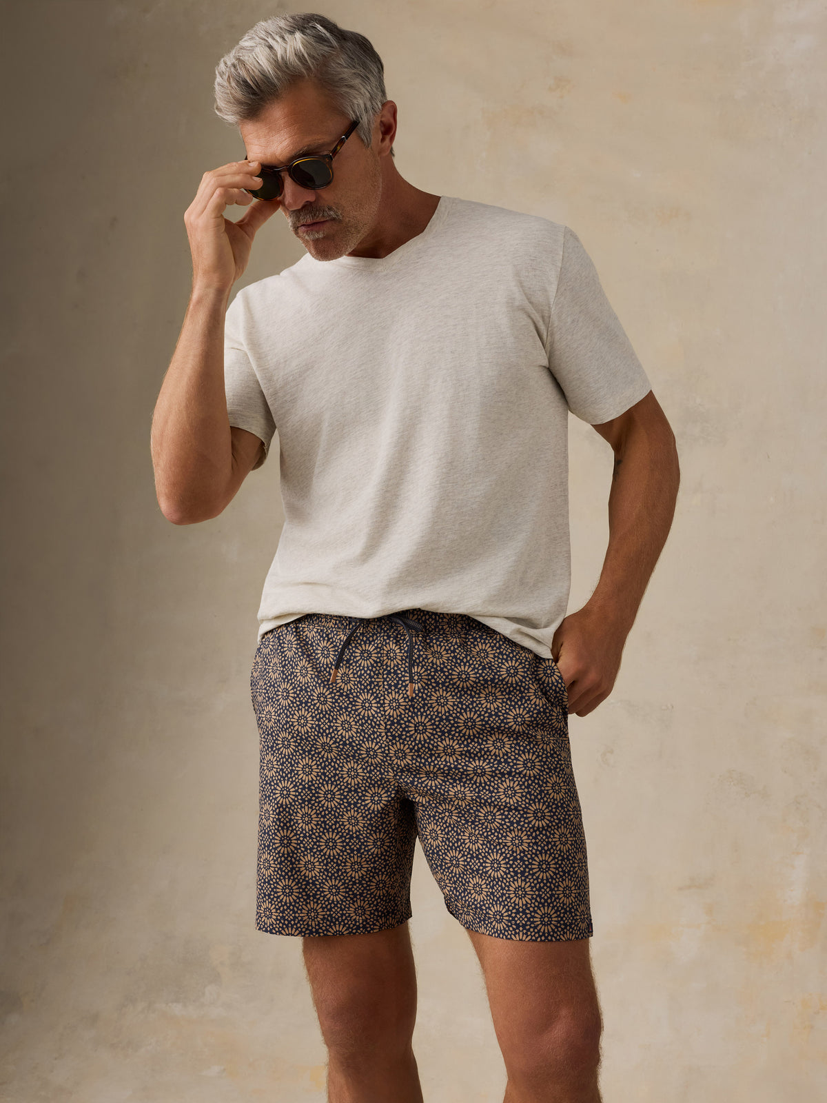 Man wearing a light colored t-shirt, brown patterned swim shorts, and sandals against a beige background, image 5 of 6
