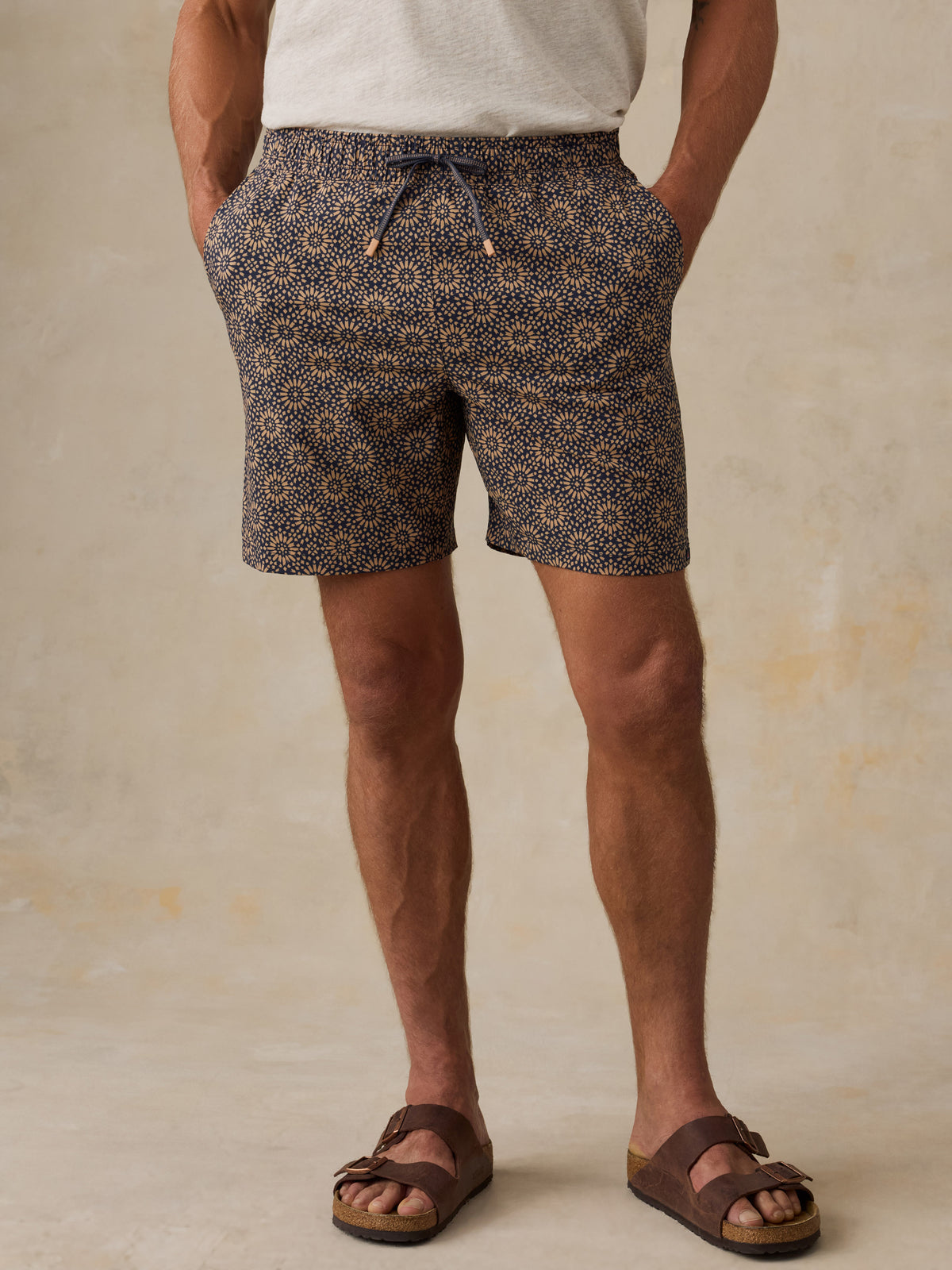 Man wearing brown patterned swim shorts and sandals against a beige background, image 1 of 6