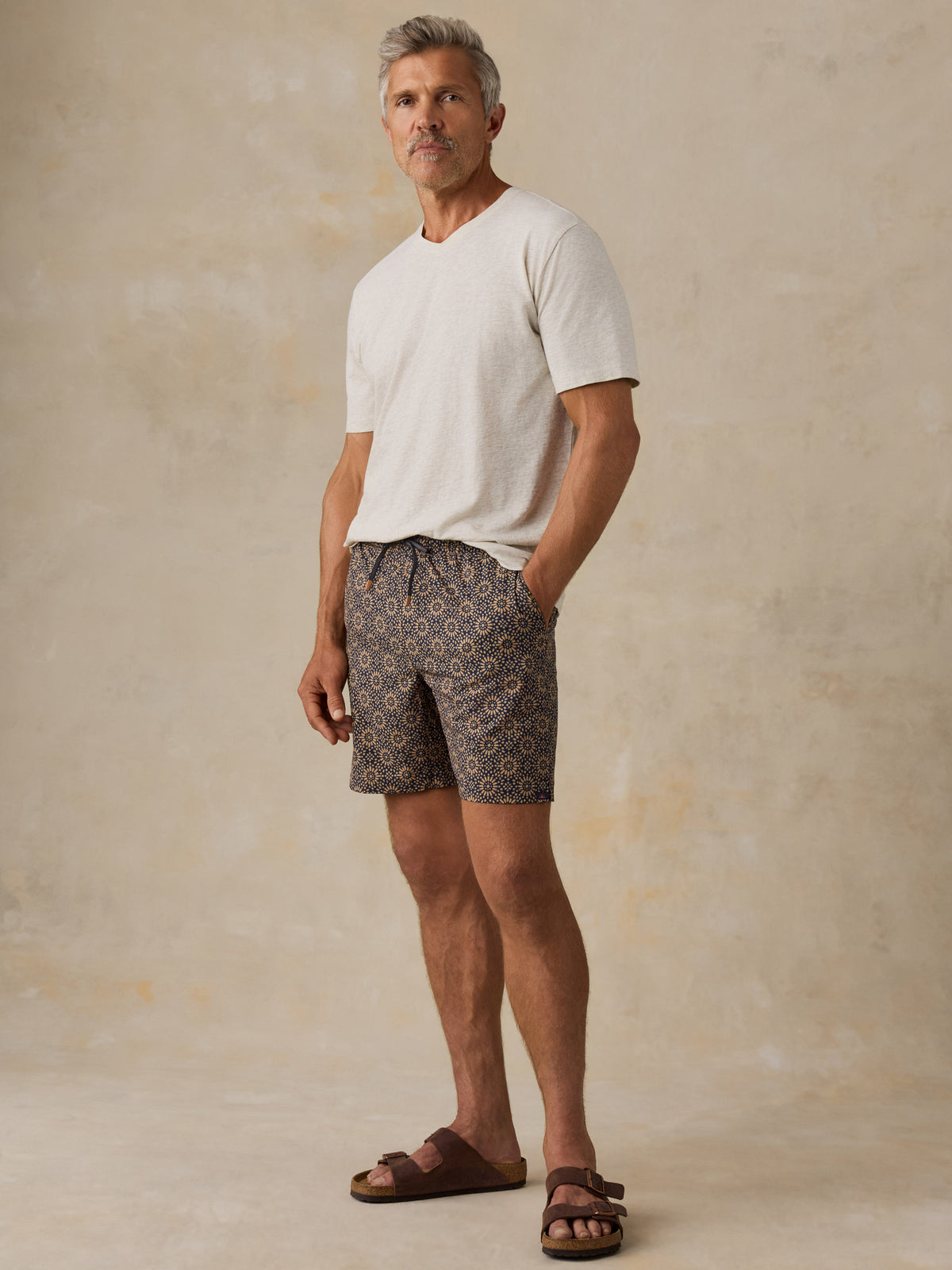 Man wearing a light colored t-shirt, brown patterned swim shorts, and sandals against a beige background, image 2 of 6