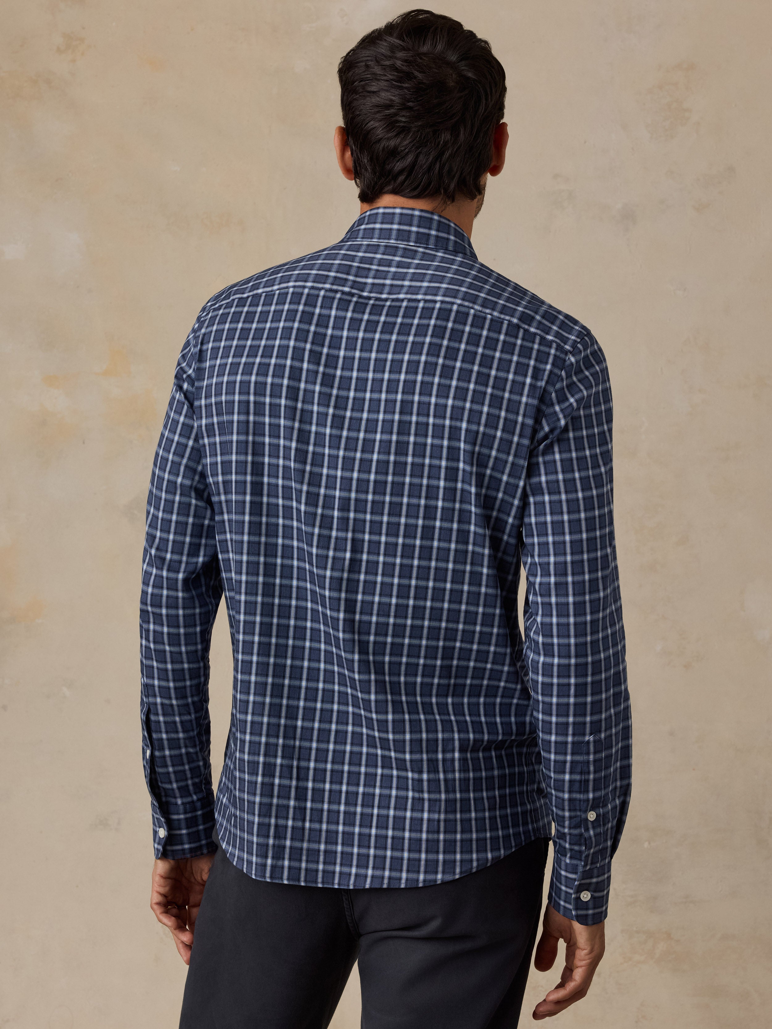 A man viewed from behind wearing a navy blue and white checkered long-sleeve shirt and dark trousers, standing against a textured beige background.