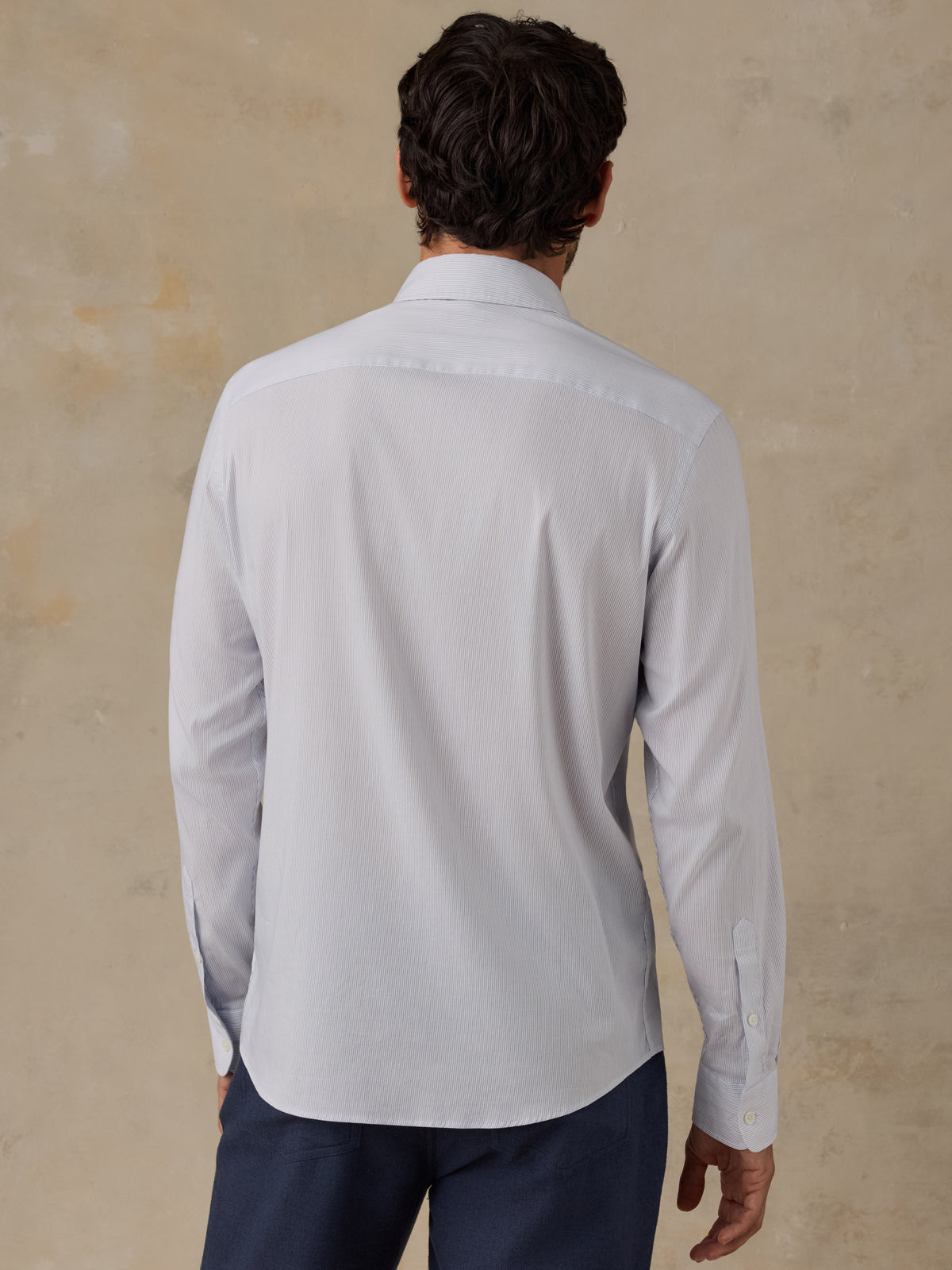A man viewed from behind wearing a light gray long-sleeve button-down shirt and navy trousers, standing against a textured beige wall., image 3 of 6