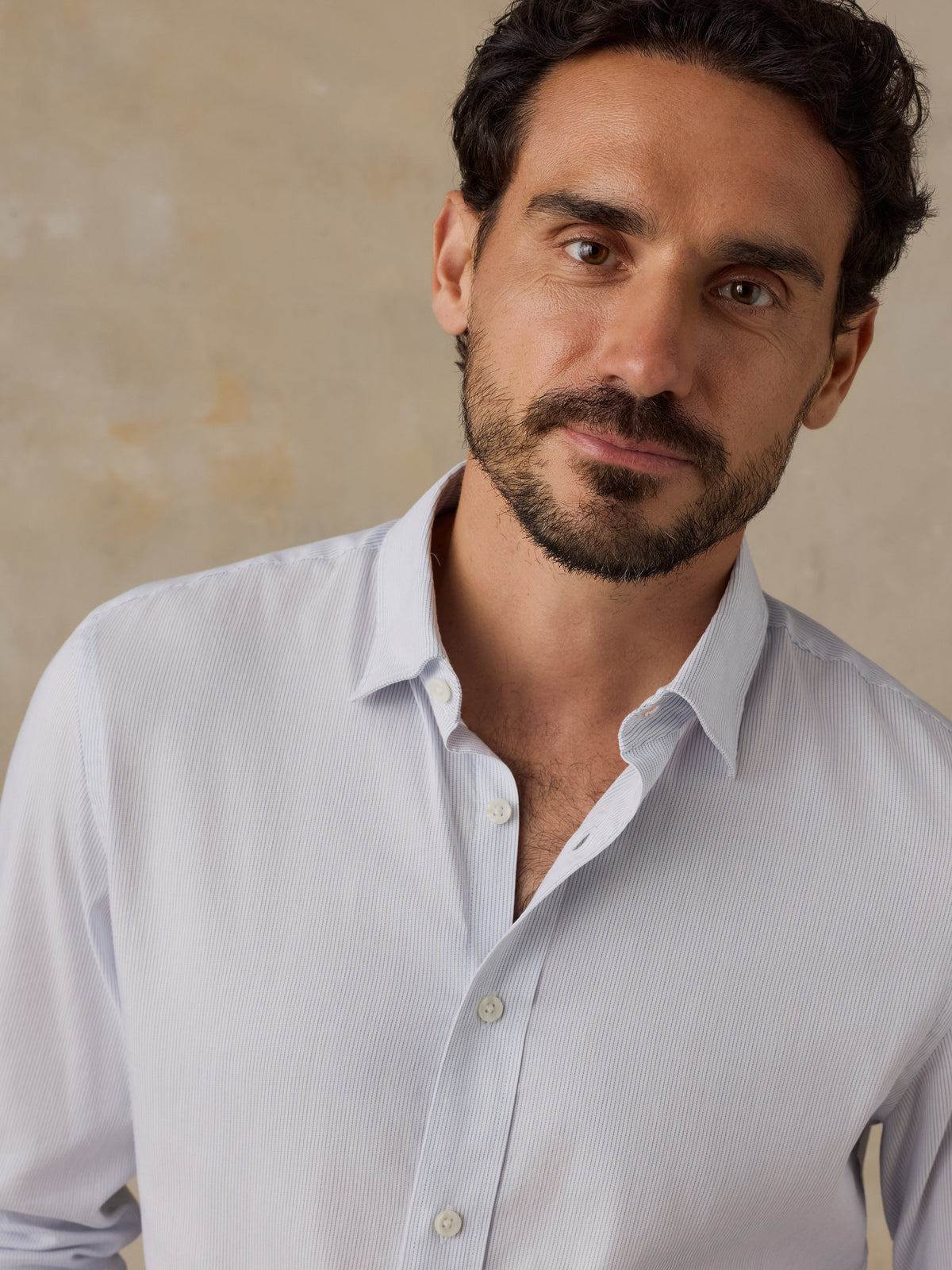A close-up portrait of a man wearing a light blue long-sleeve button-down shirt against a textured beige background., image 2 of 6