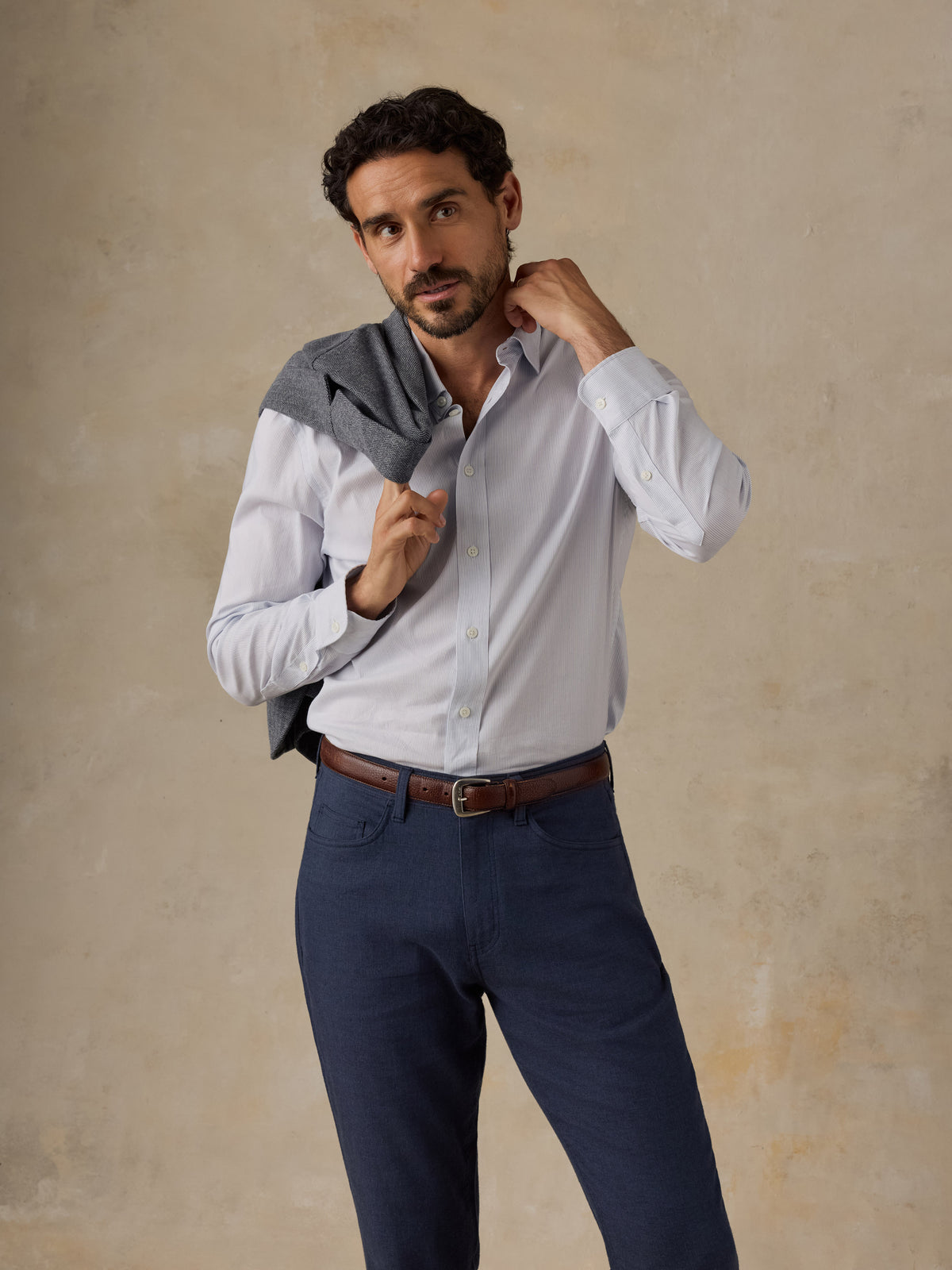 A man wearing a white long-sleeve button-down shirt and navy blue trousers, with a gray blazer draped over his shoulders, posing against a textured beige studio backdrop., image 6 of 6