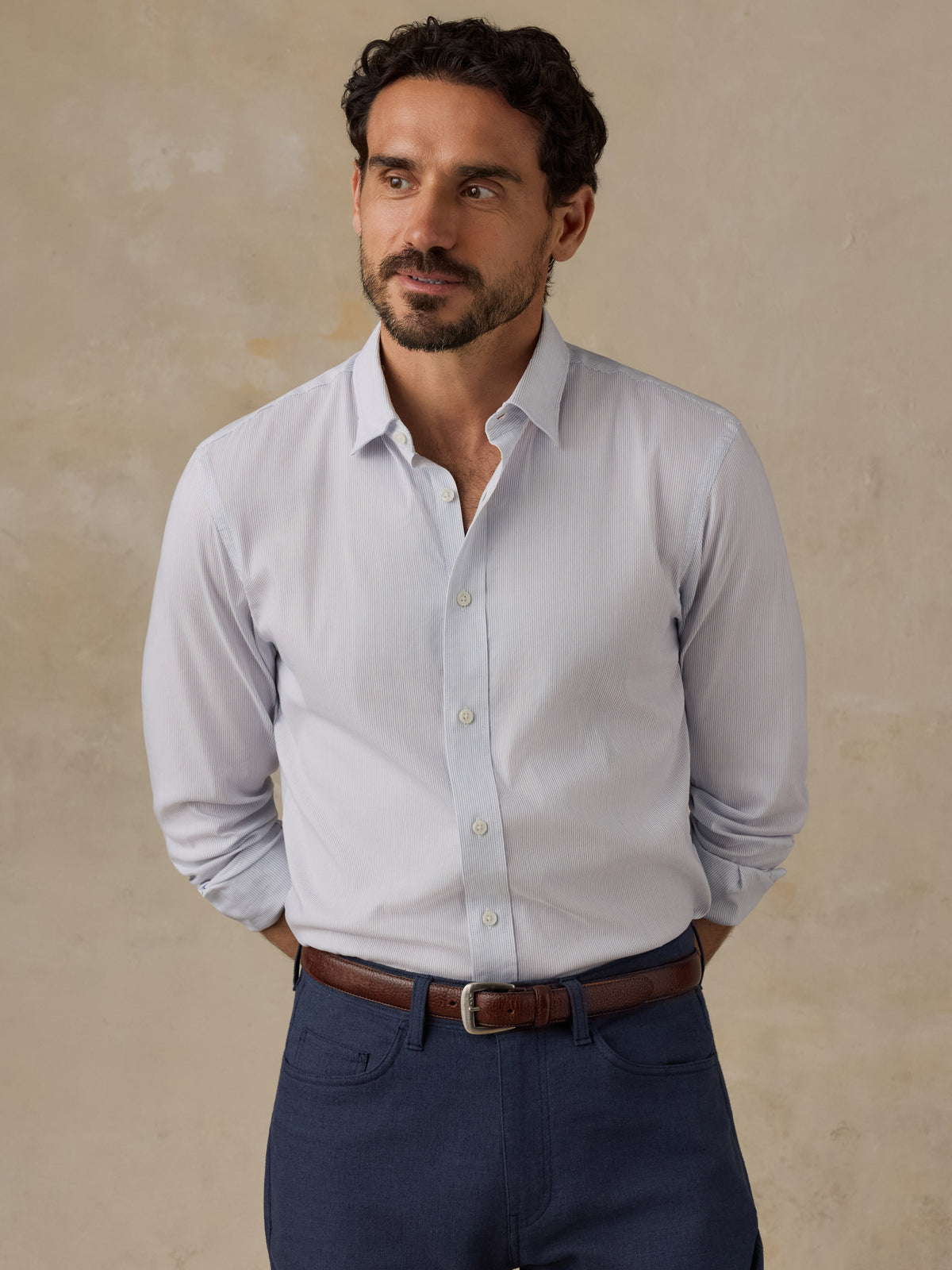 A man wearing a light blue long-sleeve button-down dress shirt, navy blue trousers, and a brown leather belt, standing against a textured beige studio backdrop., image 1 of 6