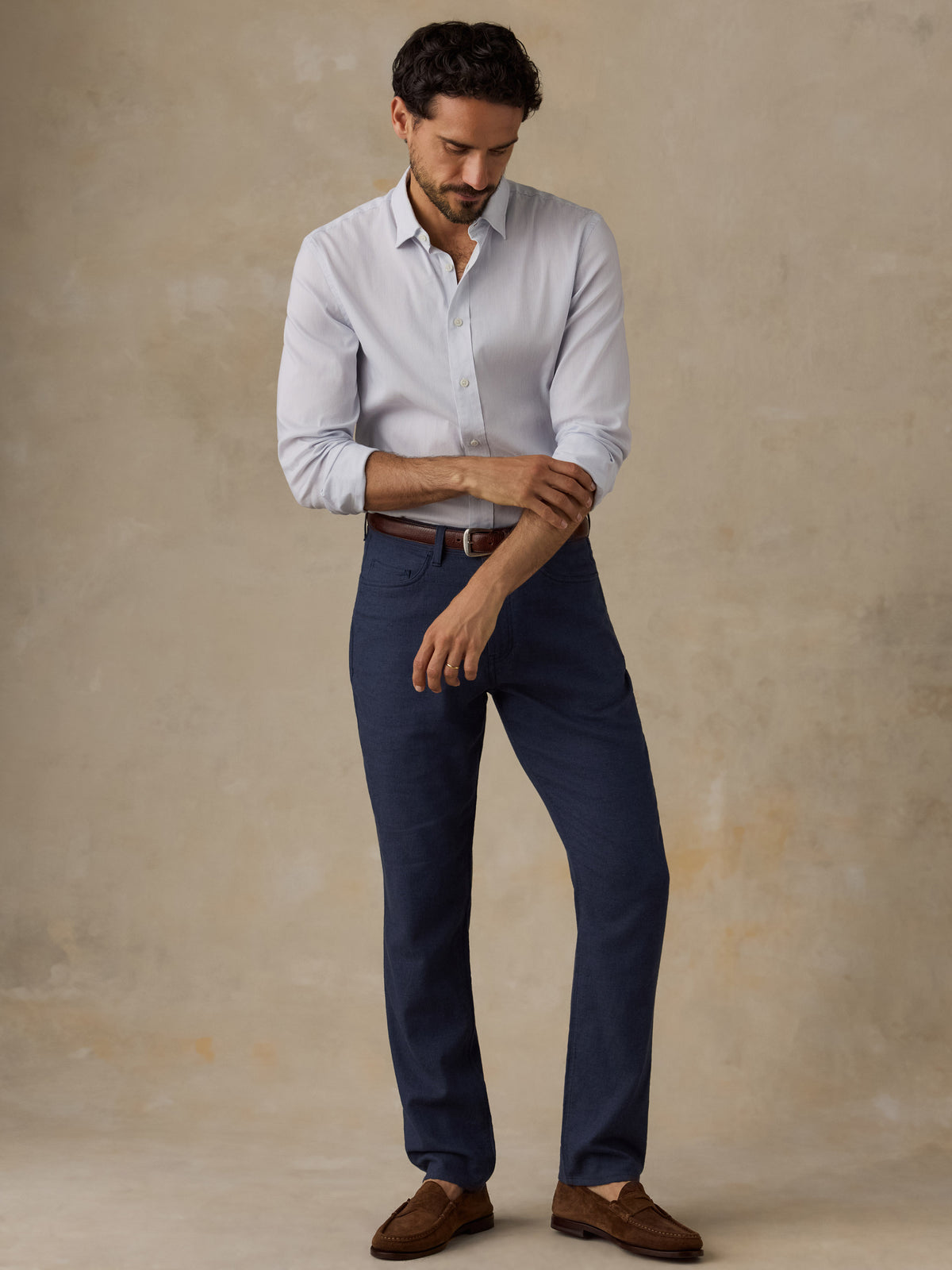 A man wearing a light blue long-sleeve button-down shirt, navy blue trousers, a brown belt, and brown leather loafers, standing against a textured beige studio backdrop., image 4 of 6