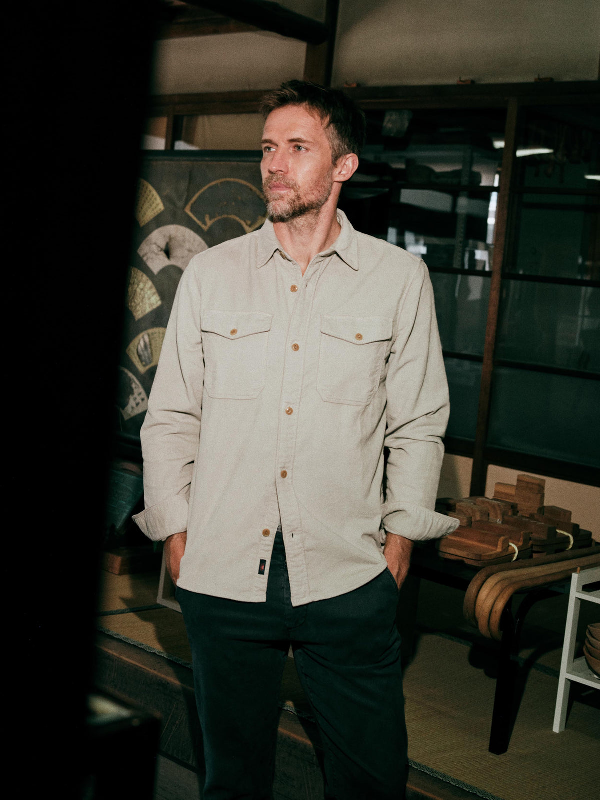 A man wearing a light beige button-down shirt with brown buttons and dark pants, image 2 of 6