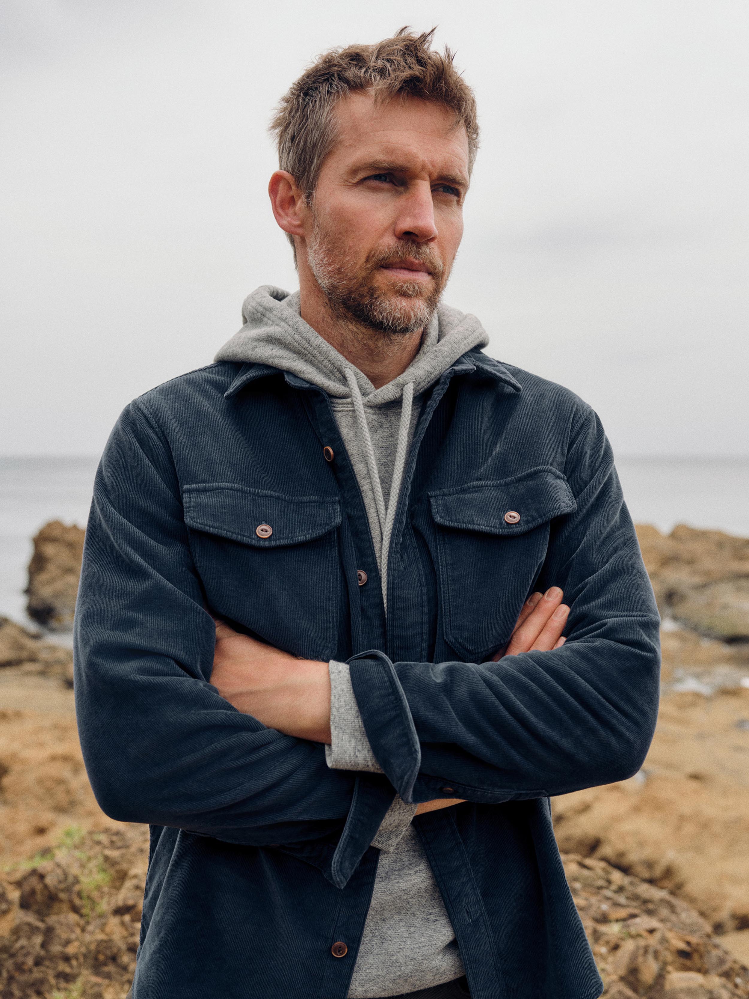 A man standing outdoors on a rocky shoreline with his arms crossed, wearing a dark navy overshirt over a light gray hoodie.