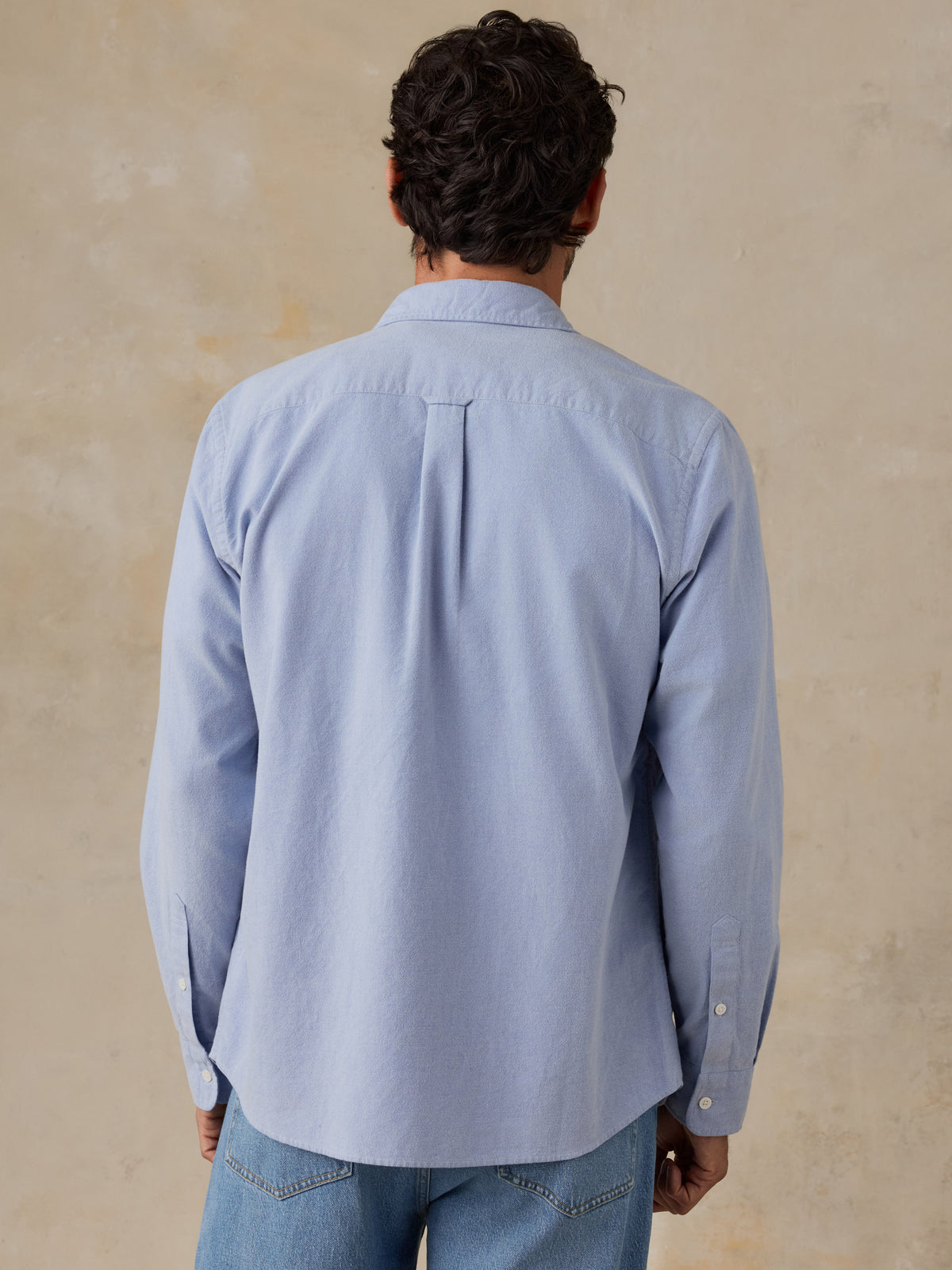 A man viewed from behind wearing a light blue long-sleeve button-up shirt and light blue jeans, standing against a textured beige wall., image 3 of 9