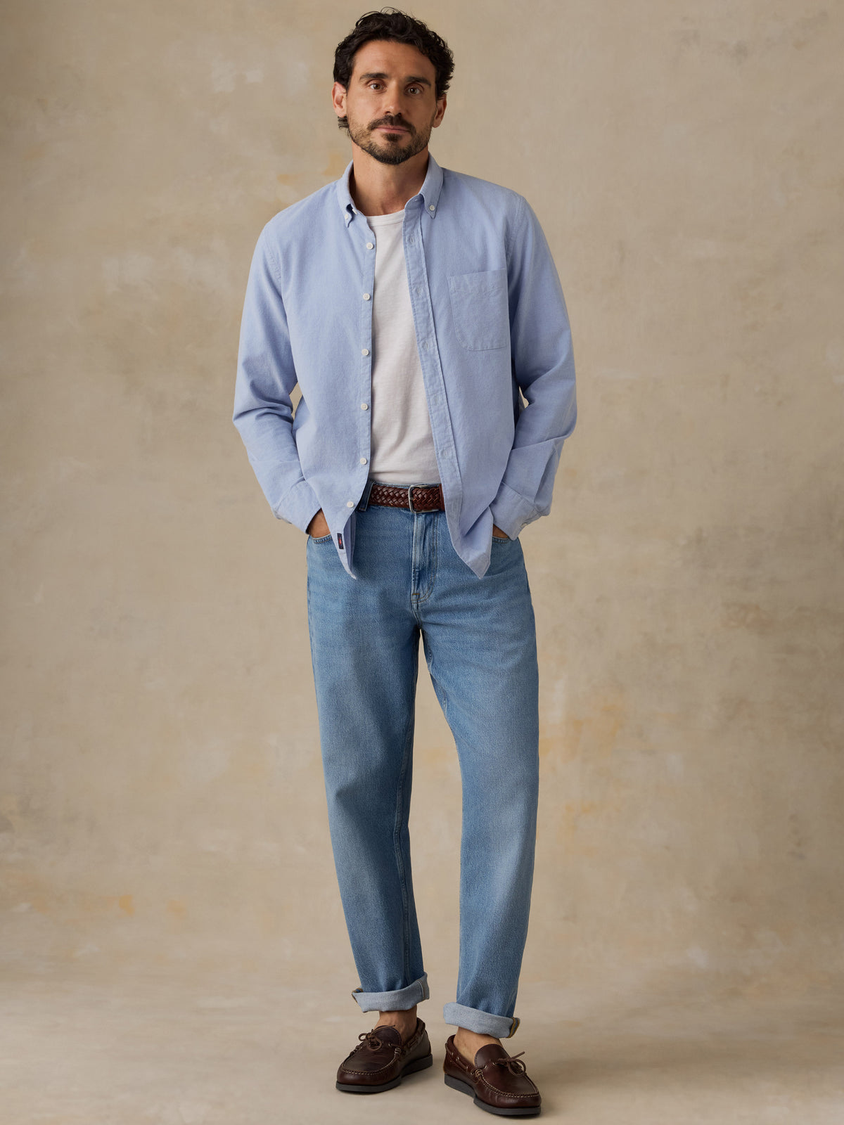 A man wearing a light blue button-down shirt over a white t-shirt, paired with light blue jeans, a brown belt, and dark brown leather boat shoes, standing against a textured beige studio backdrop., image 2 of 9