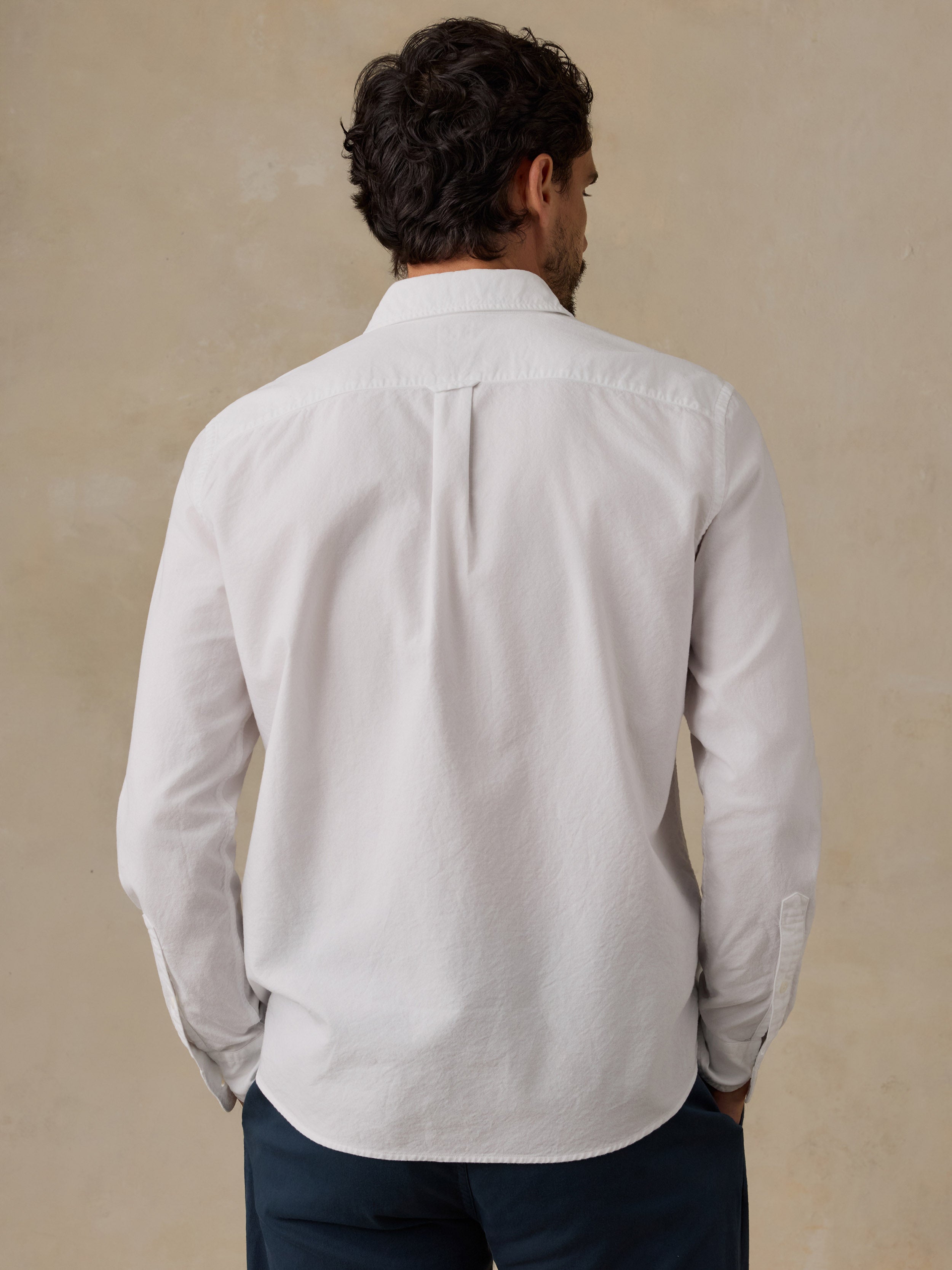 A man viewed from behind wearing a white long-sleeve button-down shirt and dark navy trousers, standing against a textured beige wall.