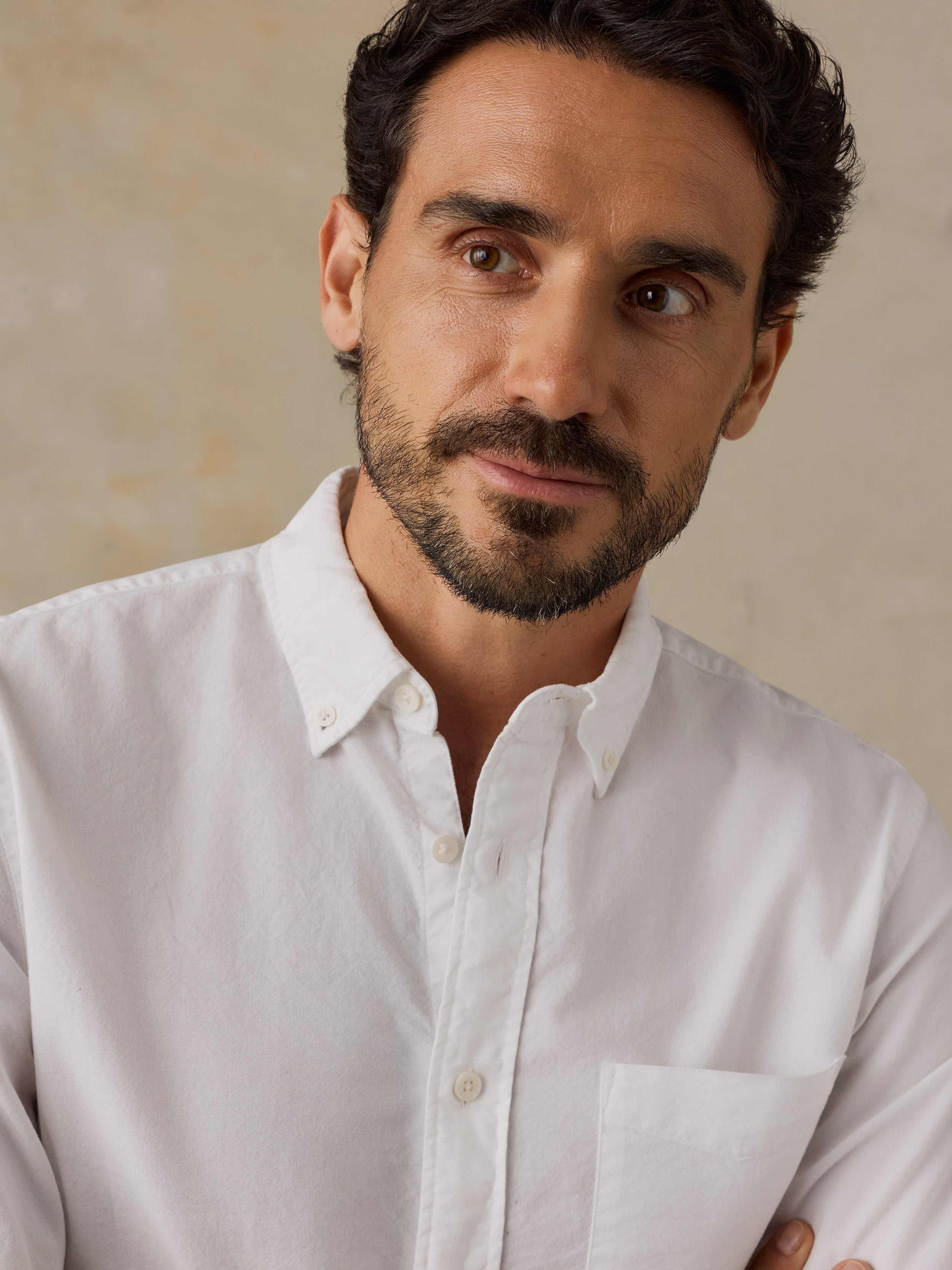A close-up portrait of a man wearing a white long-sleeve button-down shirt against a textured beige background.