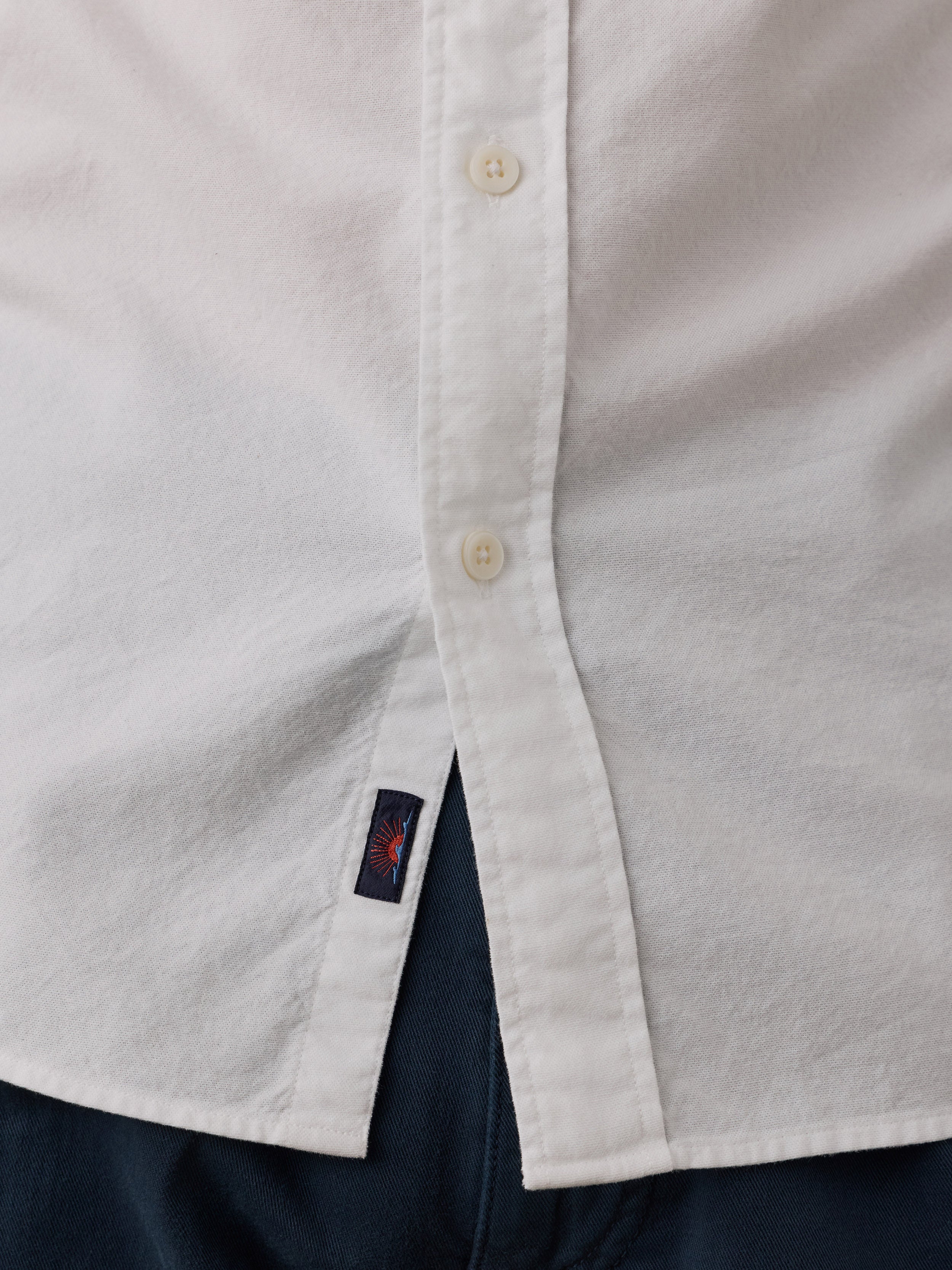 Close-up of a white button-up shirt with cream buttons and a small navy Faherty brand tag near the hem, layered over dark navy pants.