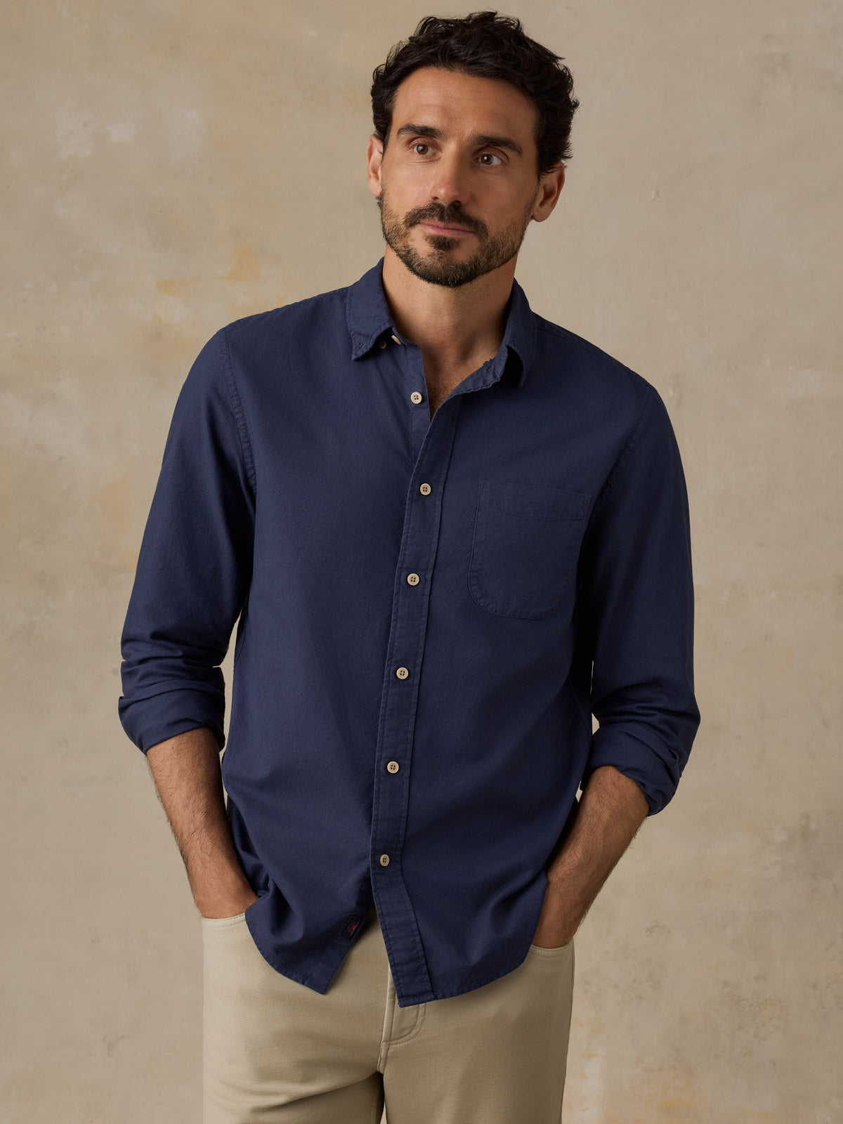 A man wearing a navy blue long-sleeve button-down shirt with rolled-up sleeves and beige trousers, standing against a textured beige wall., image 1 of 9