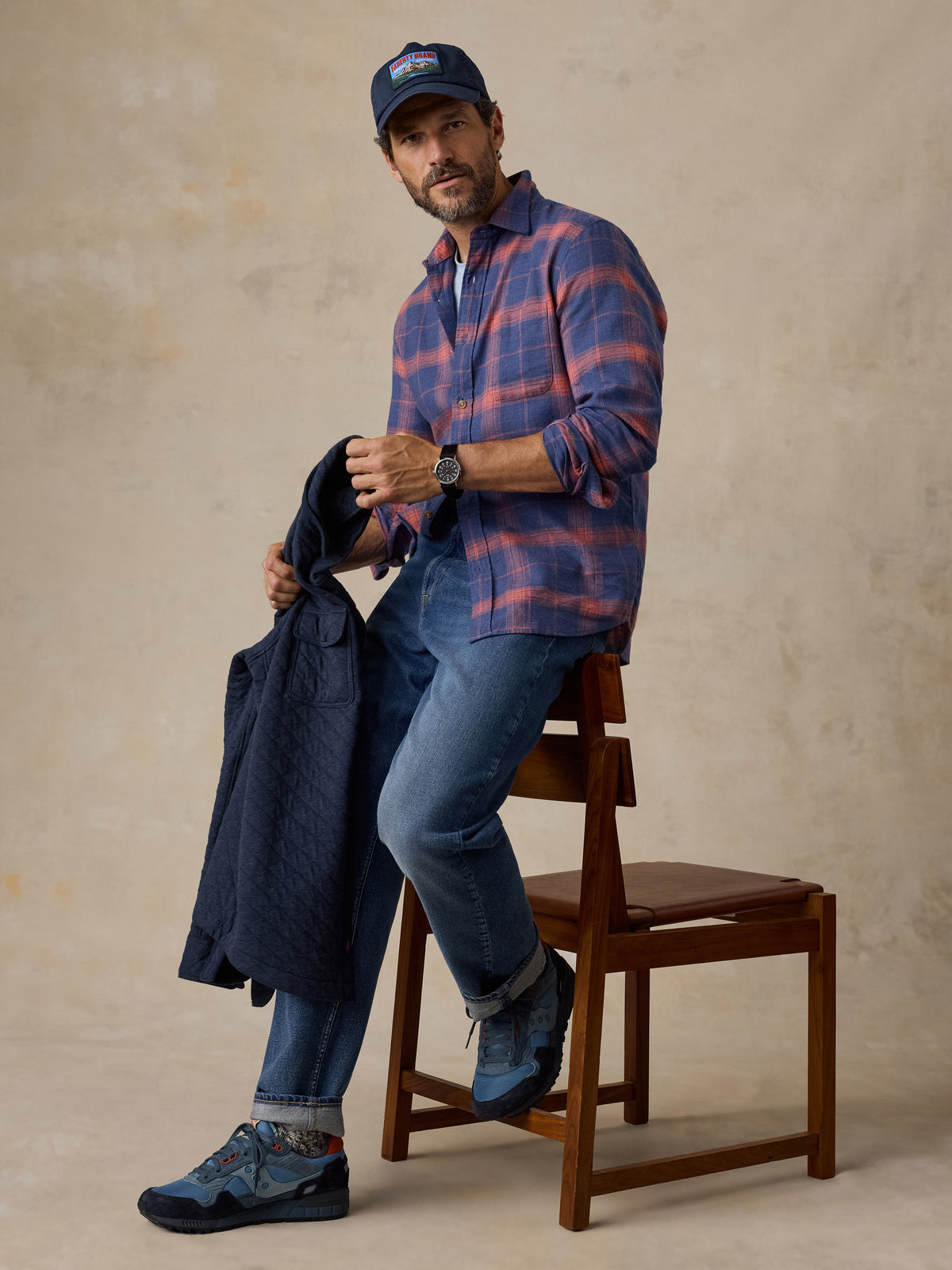 A man sitting on a chair wearing a blue plaid shirt, blue jeans, and a hat, image 2 of 5
