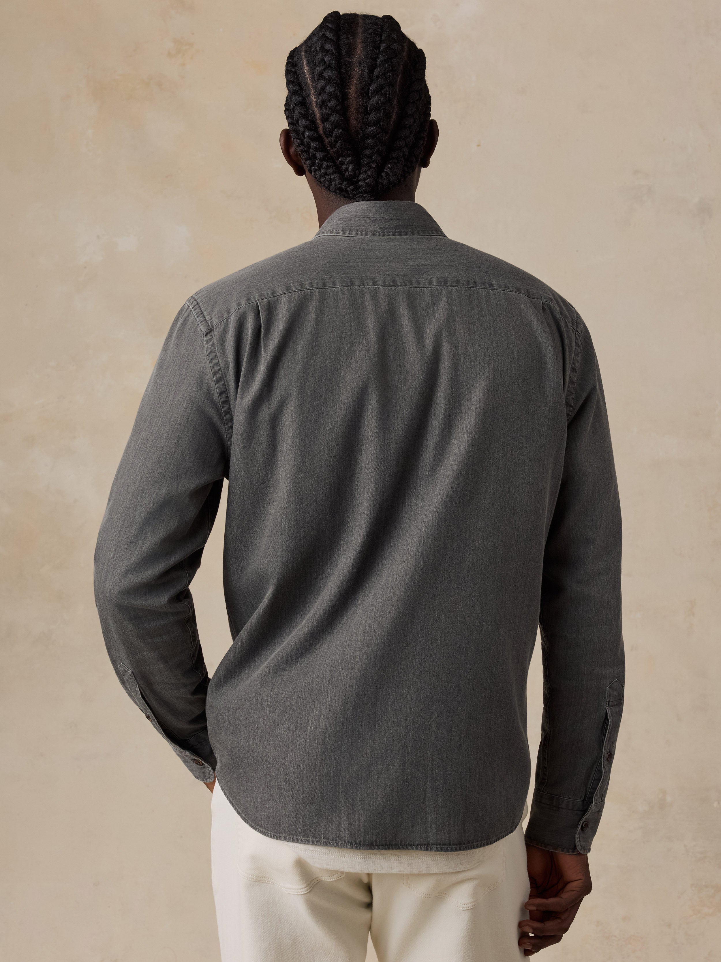 A man viewed from behind wearing a dark gray long-sleeve button-up shirt and off-white trousers, standing against a textured beige background.