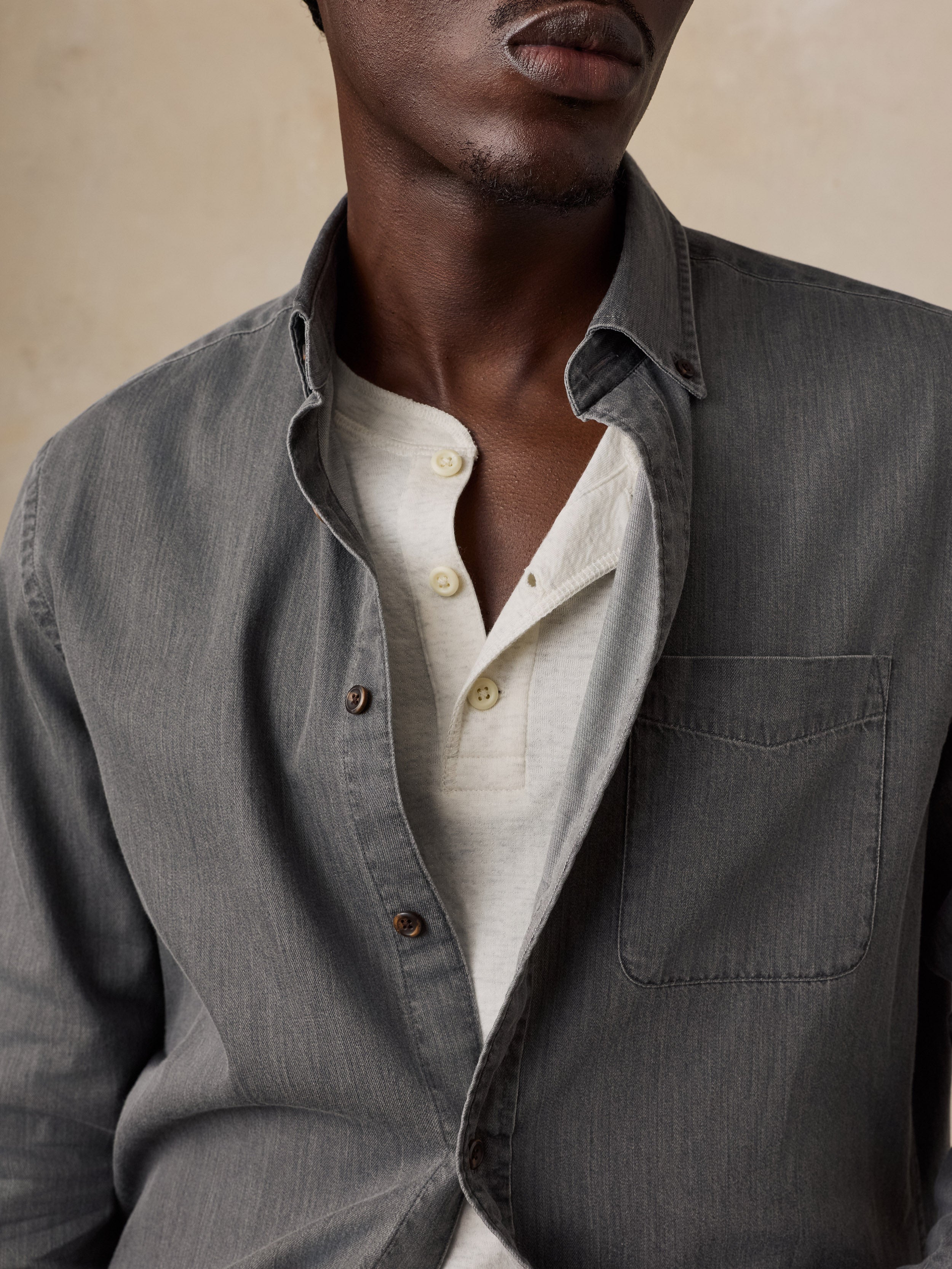 A close-up of a man wearing a dark gray textured overshirt over a light beige henley shirt, set against a neutral beige background.