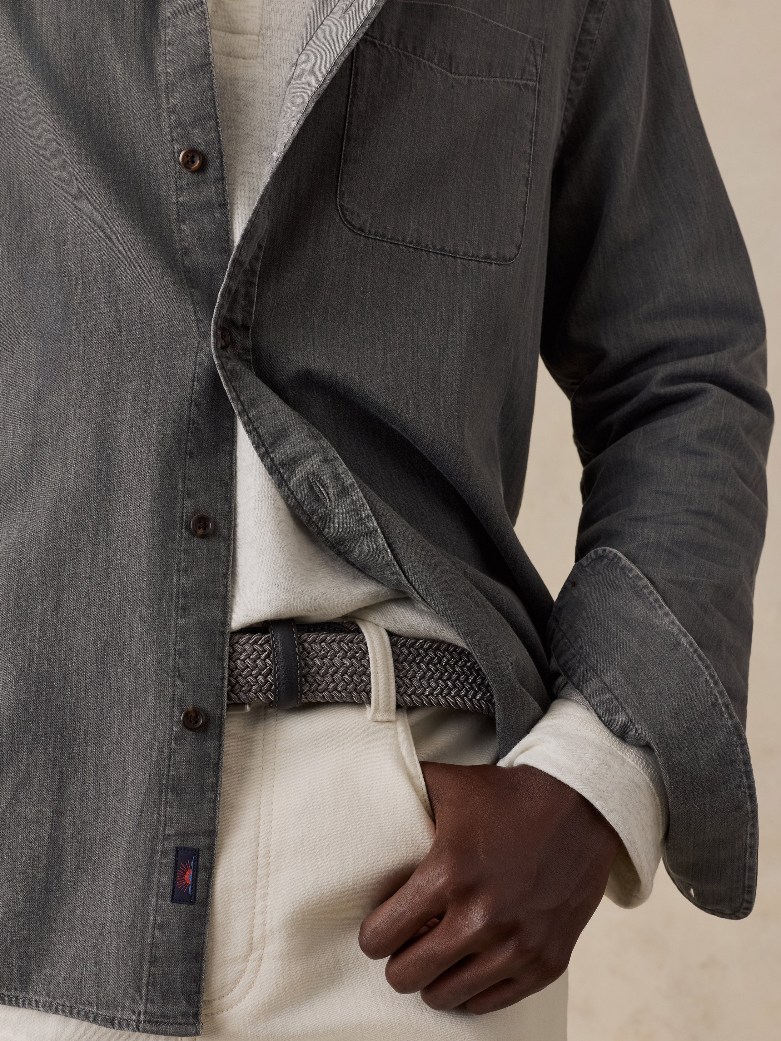 A close-up of a man wearing a dark gray chambray button-up shirt over a cream shirt, paired with a textured gray belt and cream trousers, against a neutral beige background.