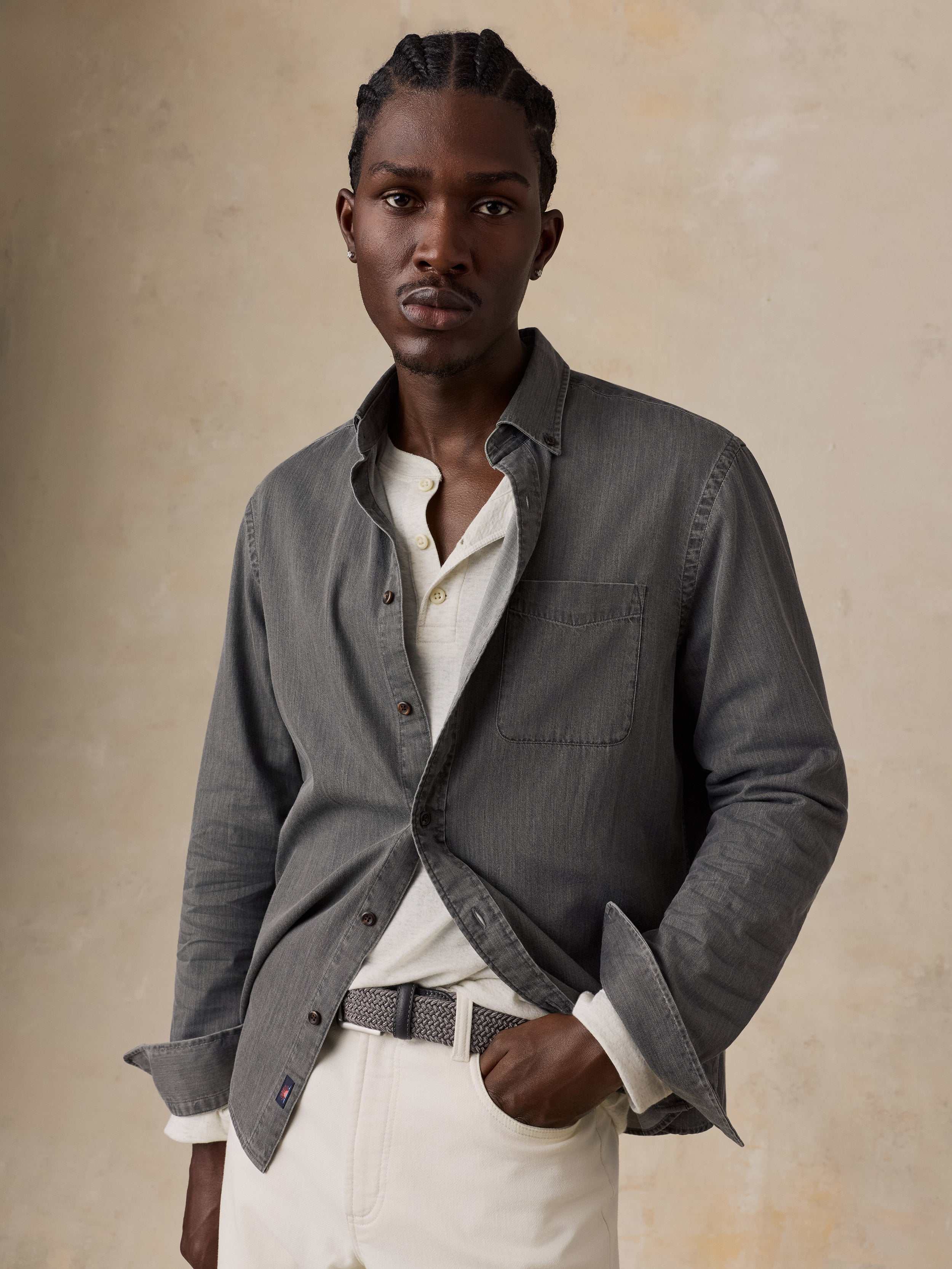 A man wearing a dark gray textured overshirt over a white collared shirt and white trousers, standing against a textured beige background.