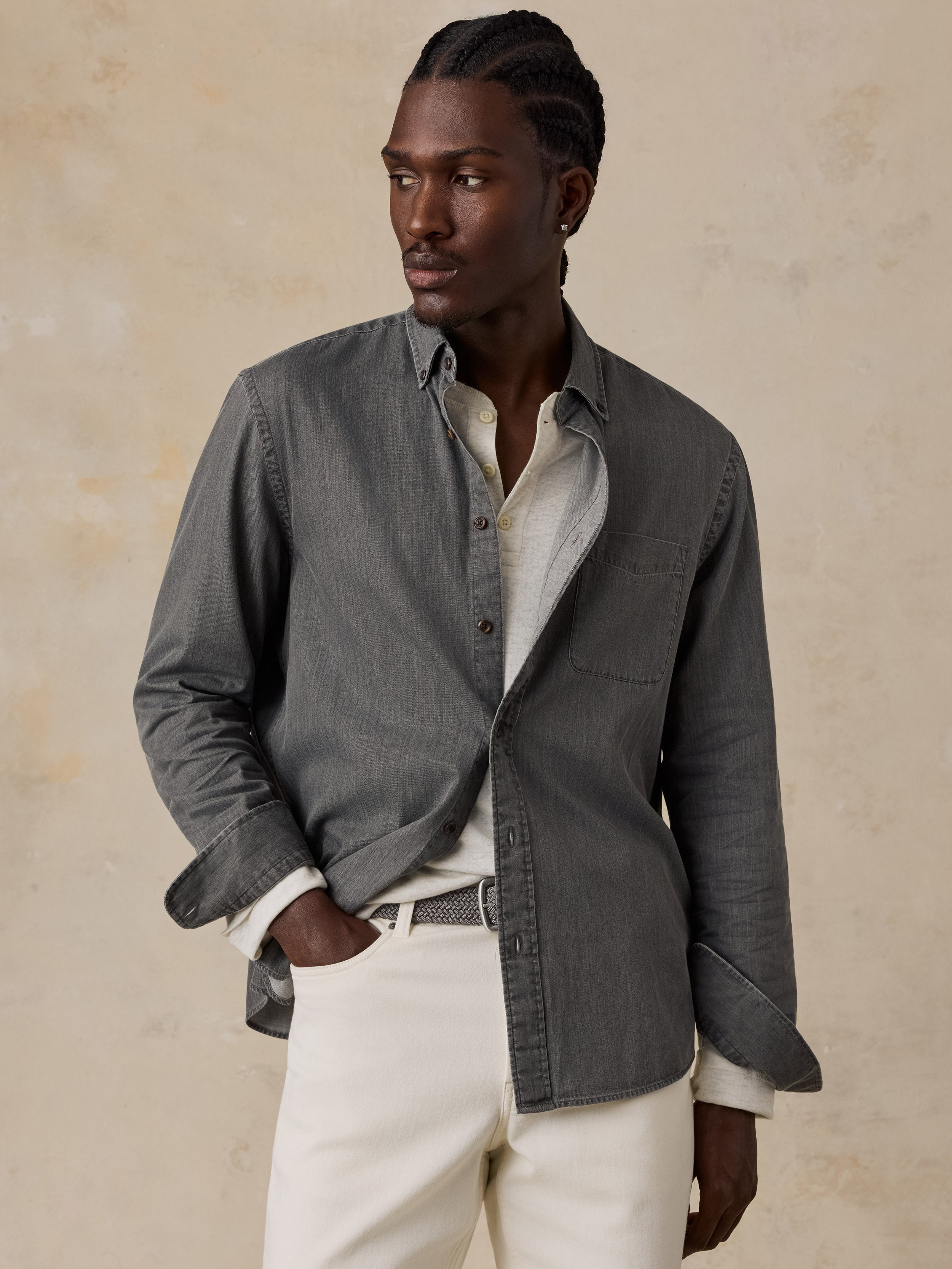 A man wearing a dark gray long-sleeve button-up shirt over a light gray tee, paired with off-white trousers, standing against a textured beige background.