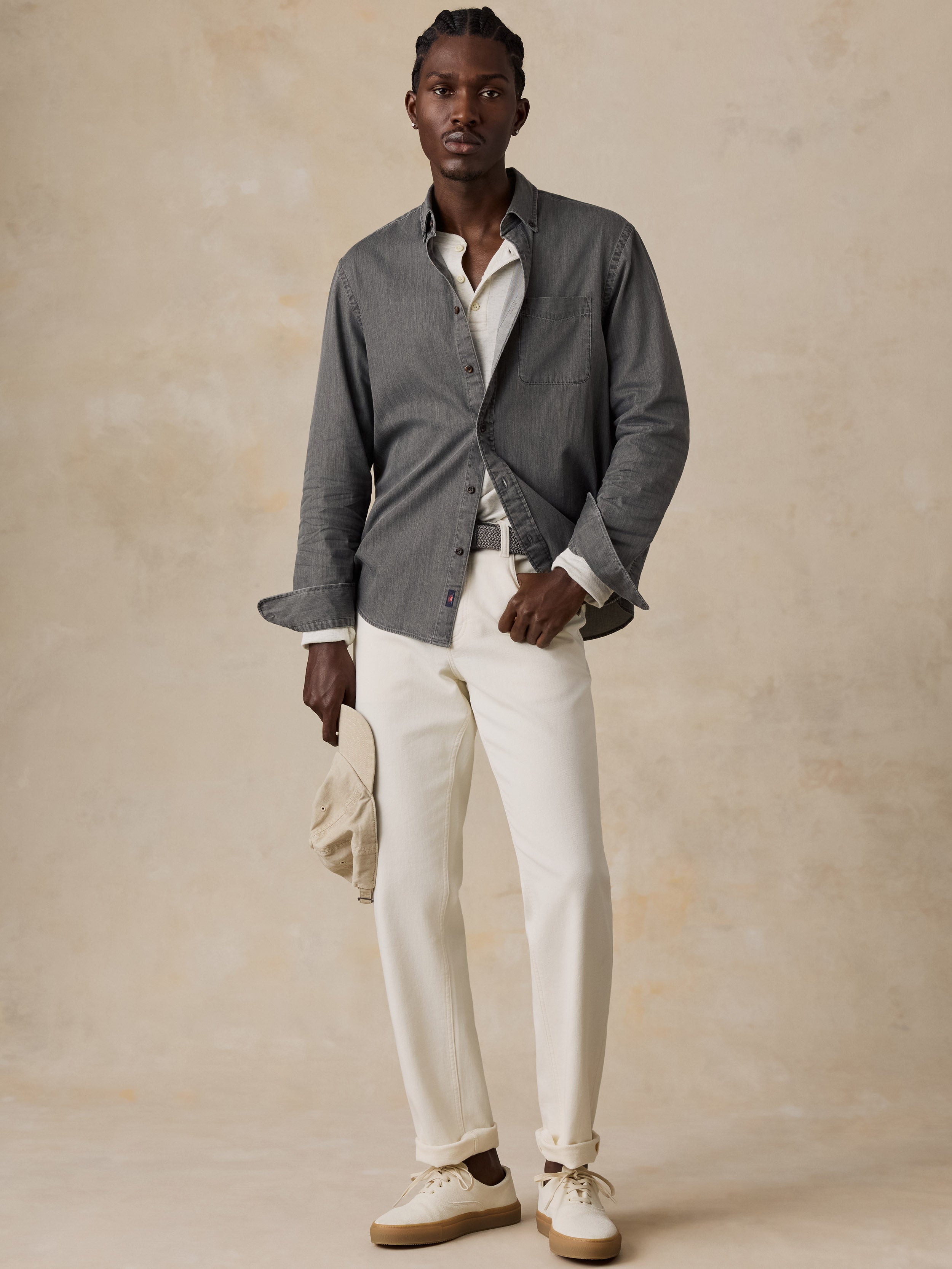 A man modeling a gray long-sleeve button-up shirt over a white collared shirt, paired with off-white trousers and beige sneakers, standing against a textured beige studio backdrop.