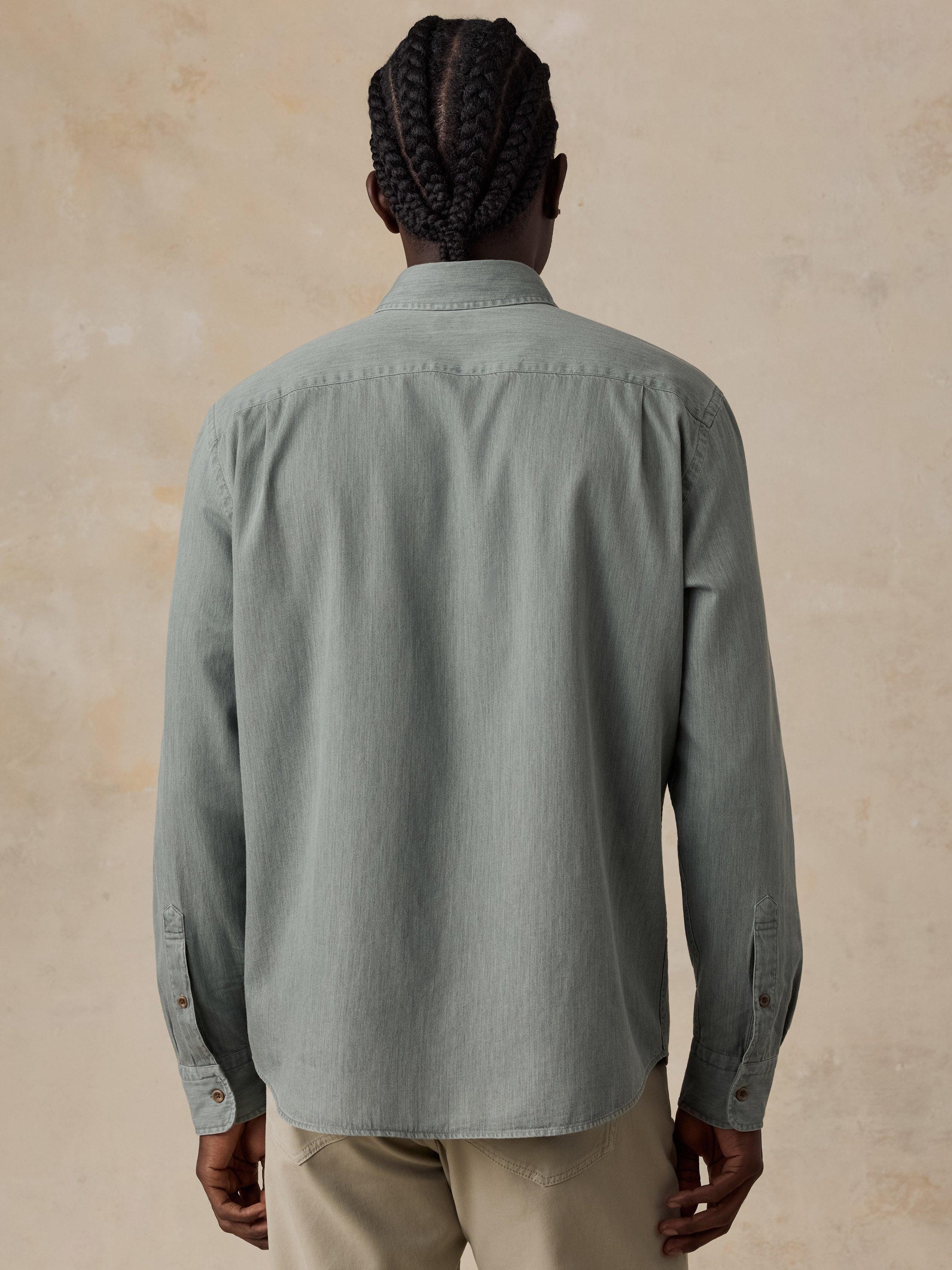 A man viewed from behind wearing a sage green long-sleeve button-down shirt and beige trousers, standing against a textured beige background.