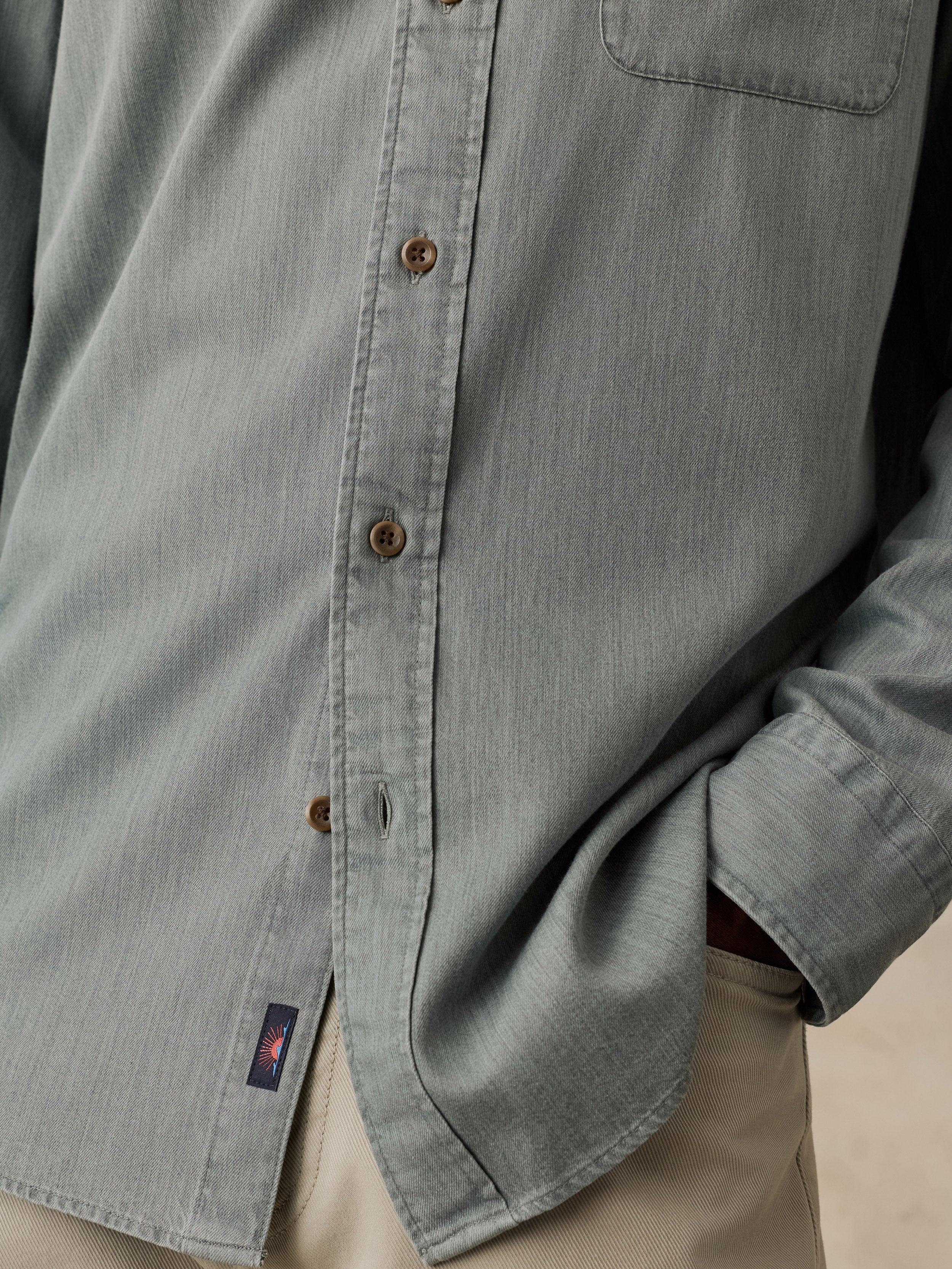Close-up of a muted sage green button-up shirt worn by a model, showing the fabric texture, brown buttons, a chest pocket, and a small logo patch near the hem.