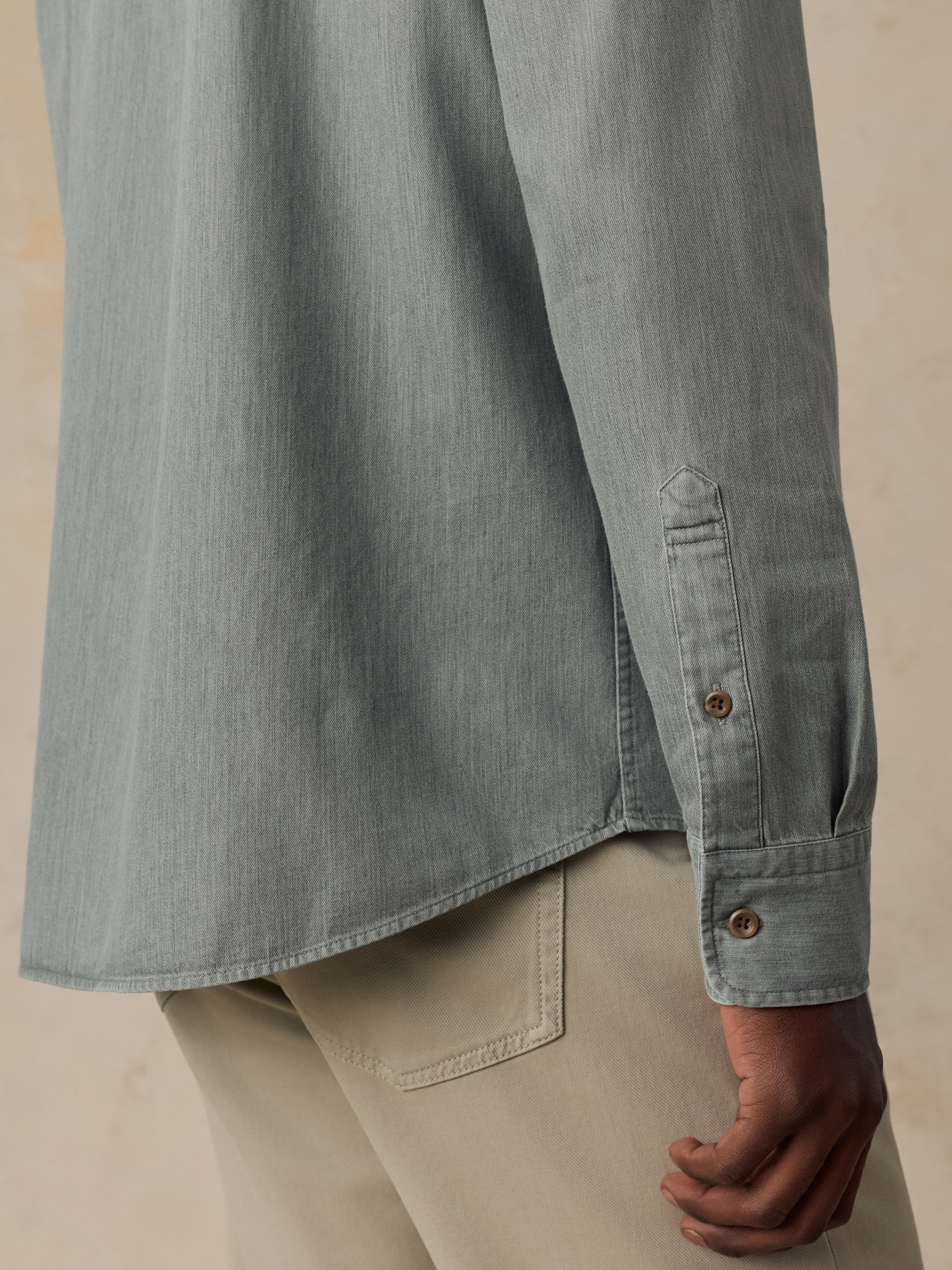 Close-up of a person wearing a muted blue-gray long-sleeve button-up shirt with beige pants, showing the fabric texture, cuff detail, and hand.